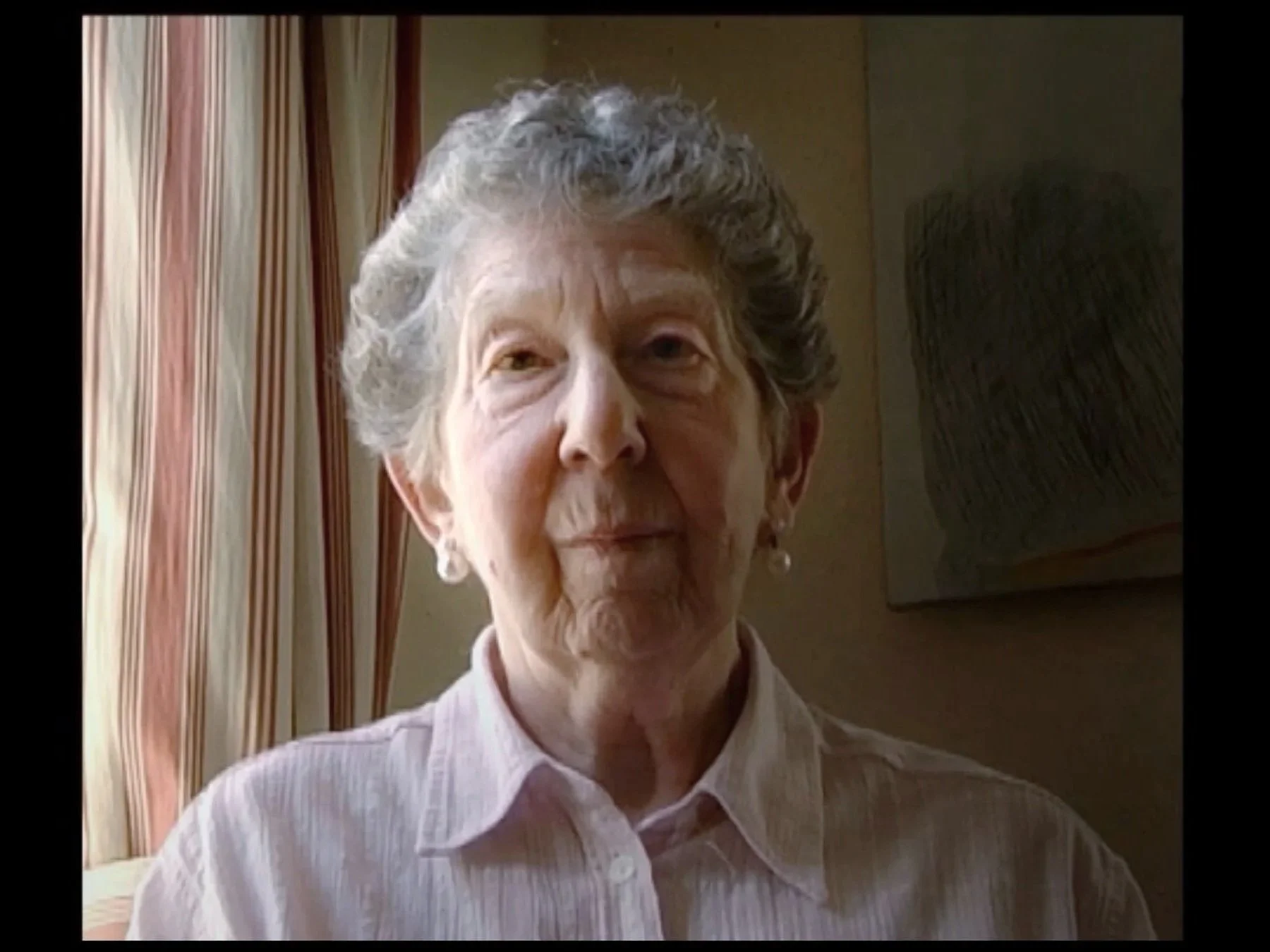 An elderly woman with short, curly gray hair wearing a pale-colored collared shirt.