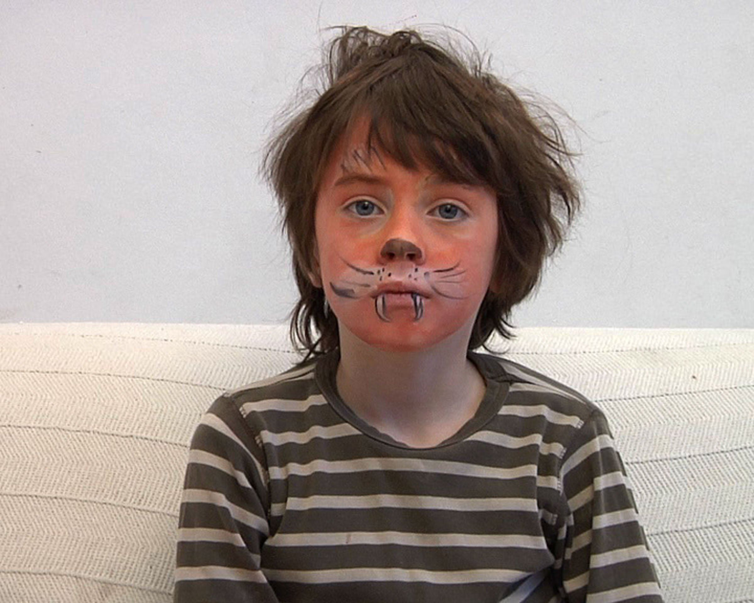 A young boy with face paint resembling a cat, wearing a striped T-shirt, sitting on a beige couch with a plain white wall in the background.