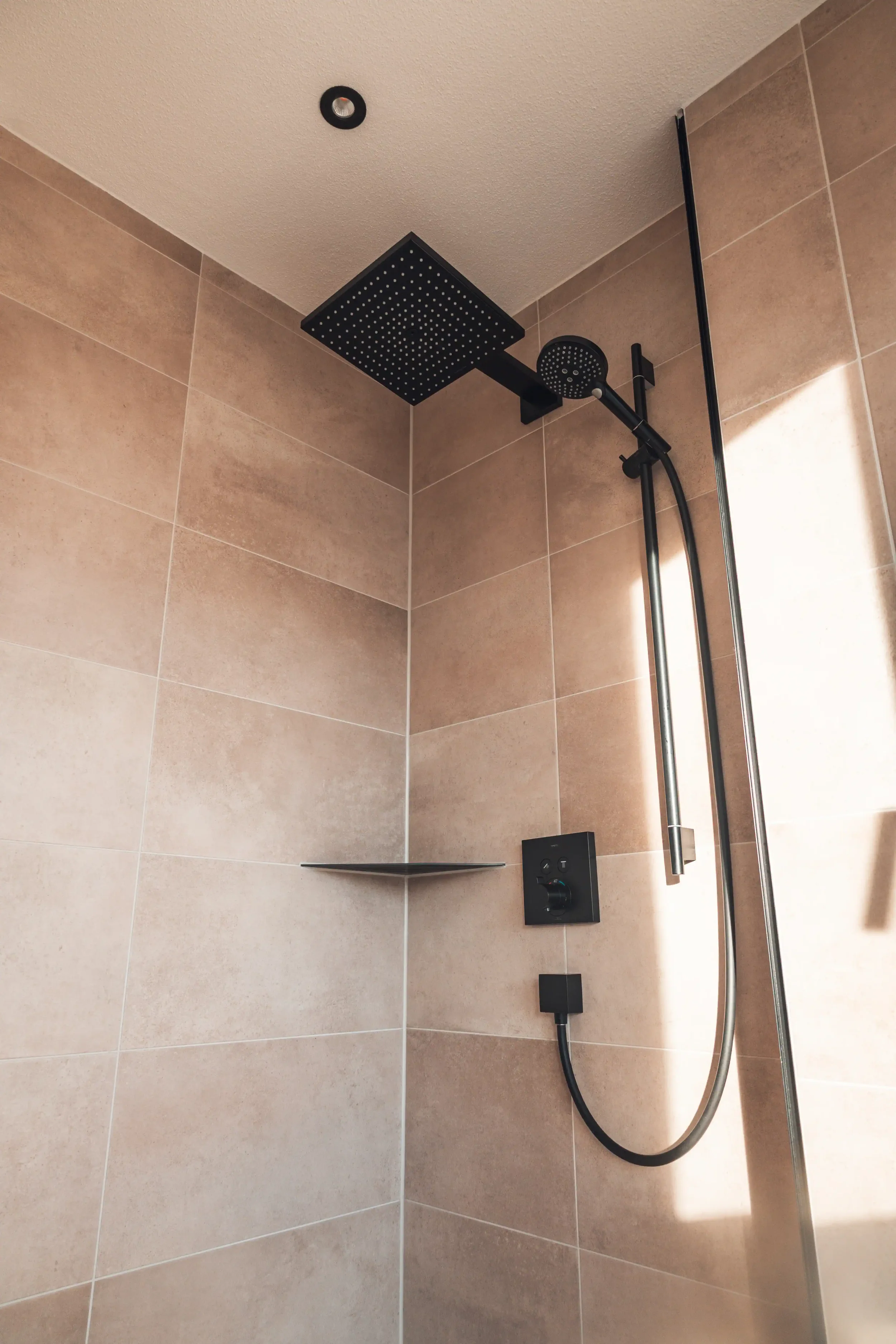 Moderne Dusche mit schwarzem Rainshower Kopf und Handbrause, beige Fliesen, höhenverstellbare Duschstange, dunkle Duschsteuerung, kleiner Ablagekorb.