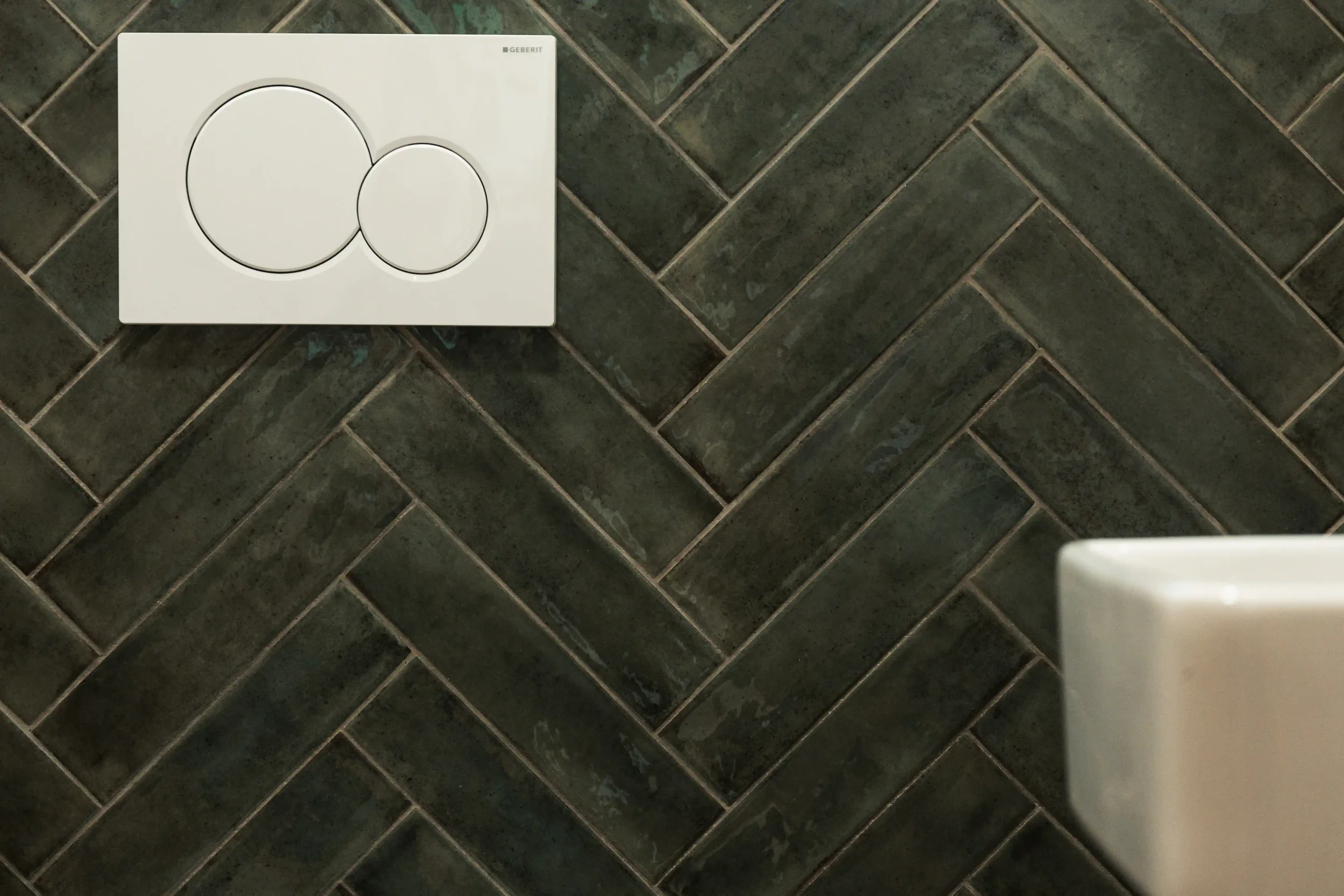 Wand mit grüner Fliesenmosaik und weißem Toilettenspülknopf
