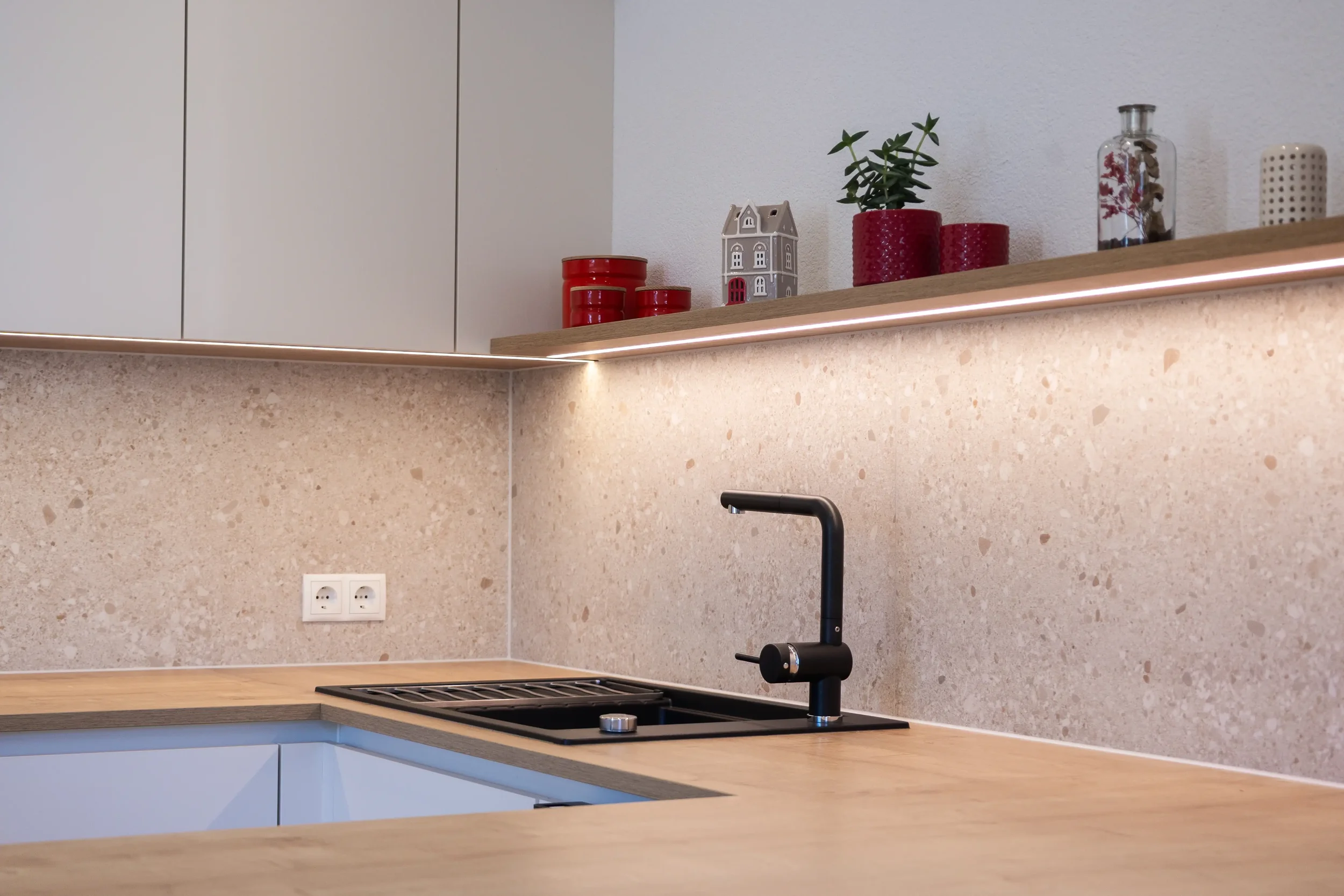 Moderne Küche mit schwarzen Wasserhahn, Holzarbeitsplatte, beige Mosaikstein-Wand, weißen oberen Schränken, dekorativen roten und grauen Vasen und Pflanzen auf offener Holzregal.