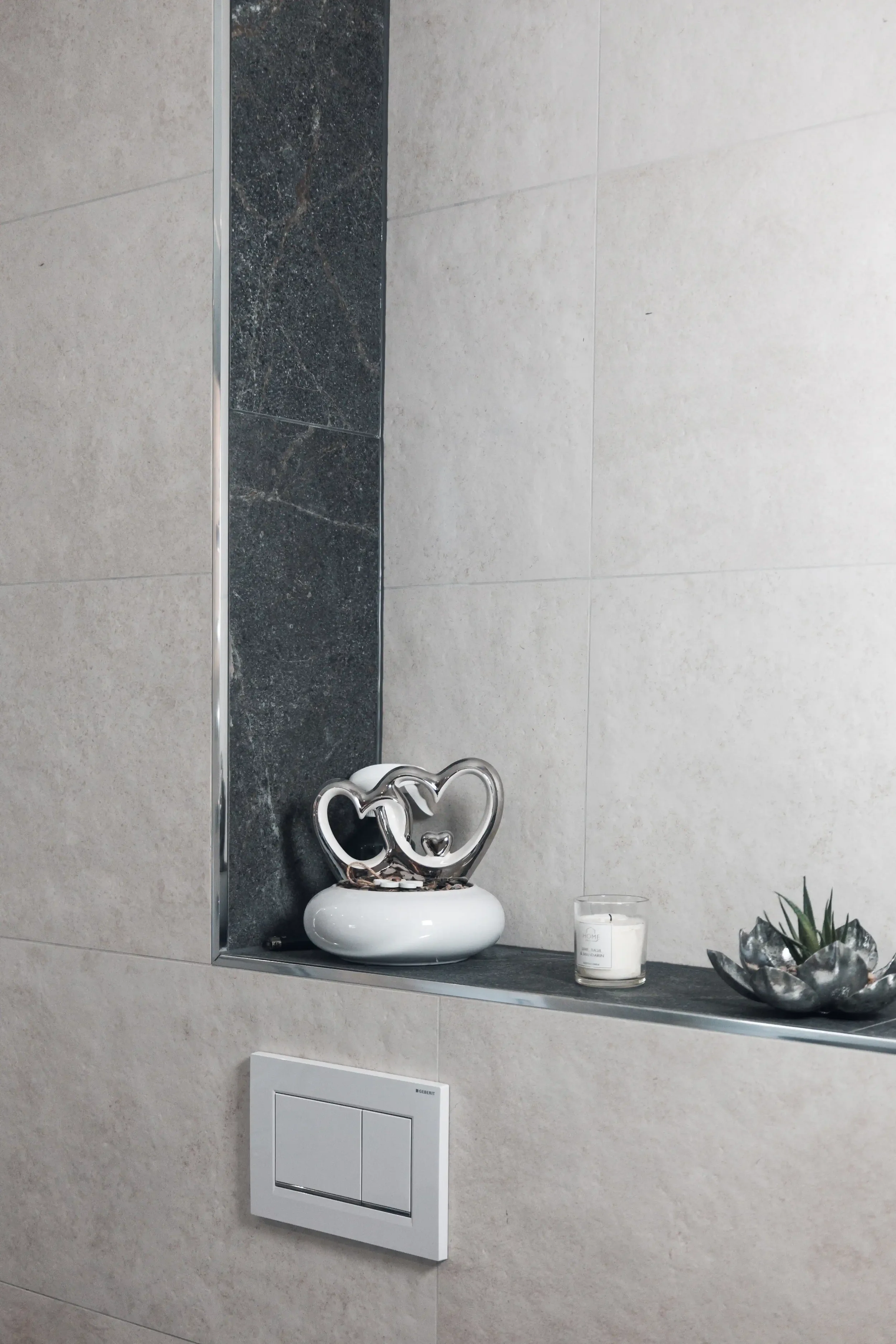 Dekorativer Kerzenhalter mit zwei Herzen, eine kleine Kerze in einem Glas, eine Sukkulente in einer silberfarbenen Schale, alles auf einer Keramiknische in einer modernen Toilette oder Bad