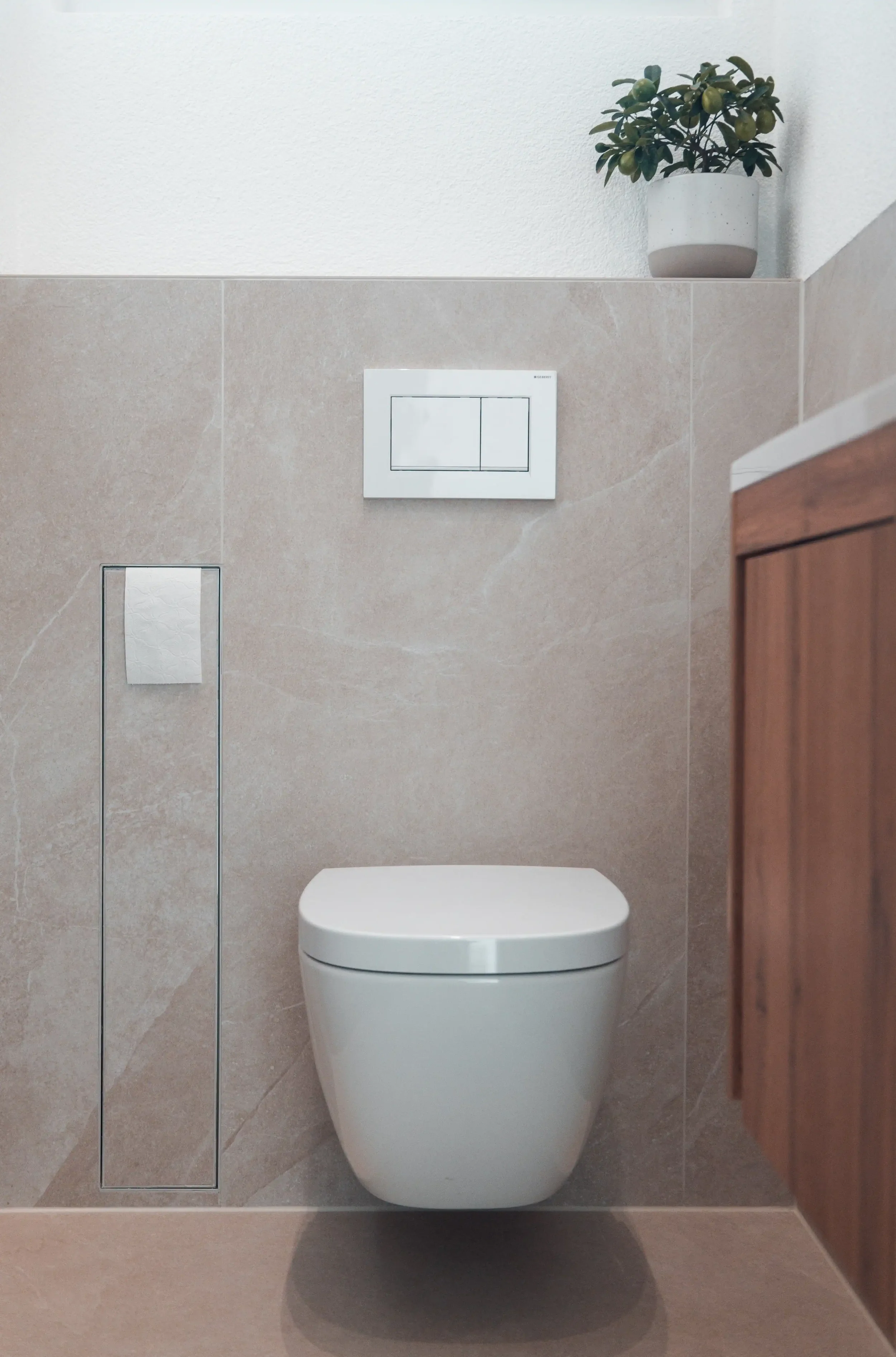 Moderne Toilette in einem Badezimmer mit beige Fliesen, einem Holzschrank und einer Pflanze in einem weißen Top