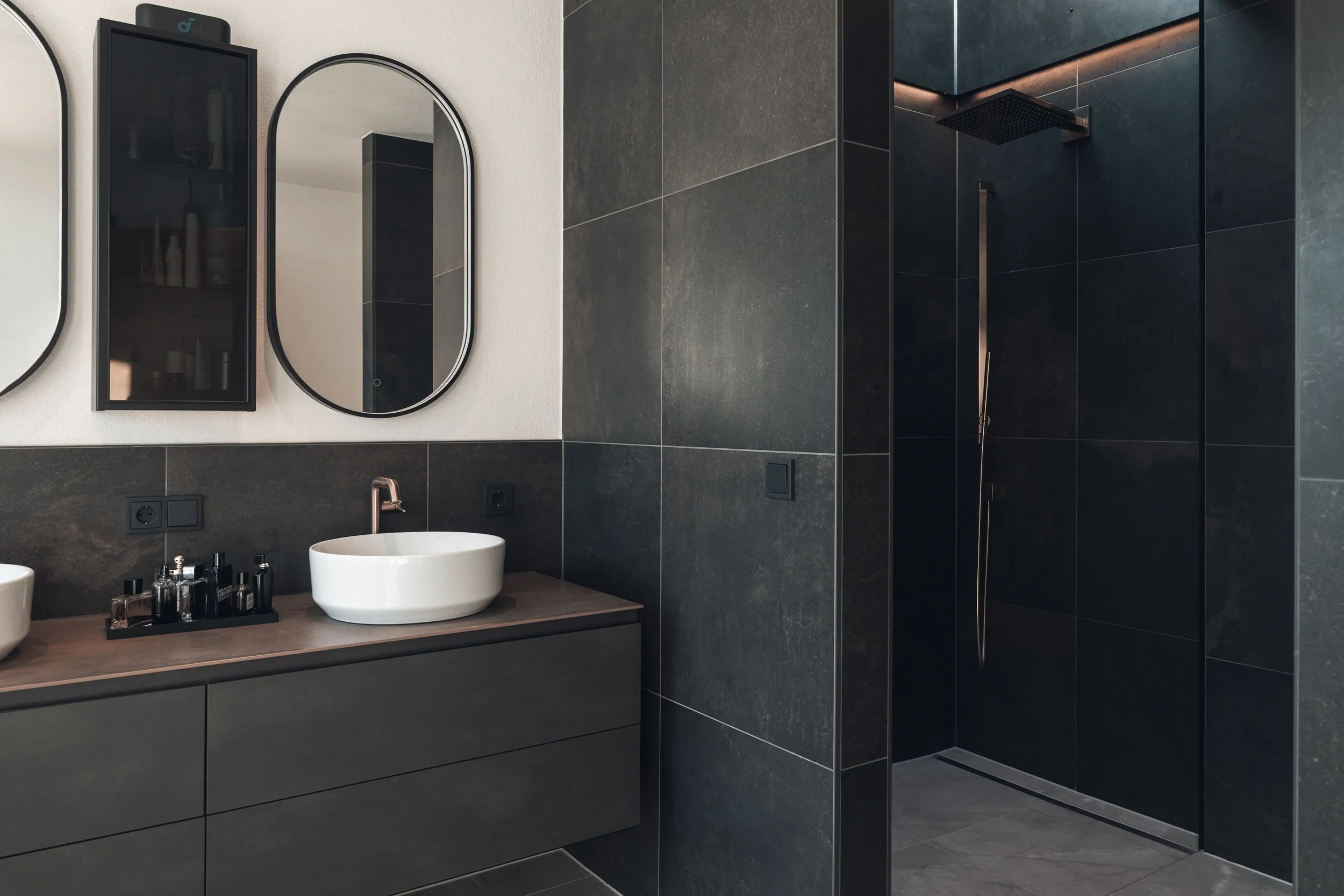 Moderner Badezimmerbereich mit schwarzen Fliesen, Doppelspiegel, Waschbecken und begehbarem Duschbereich