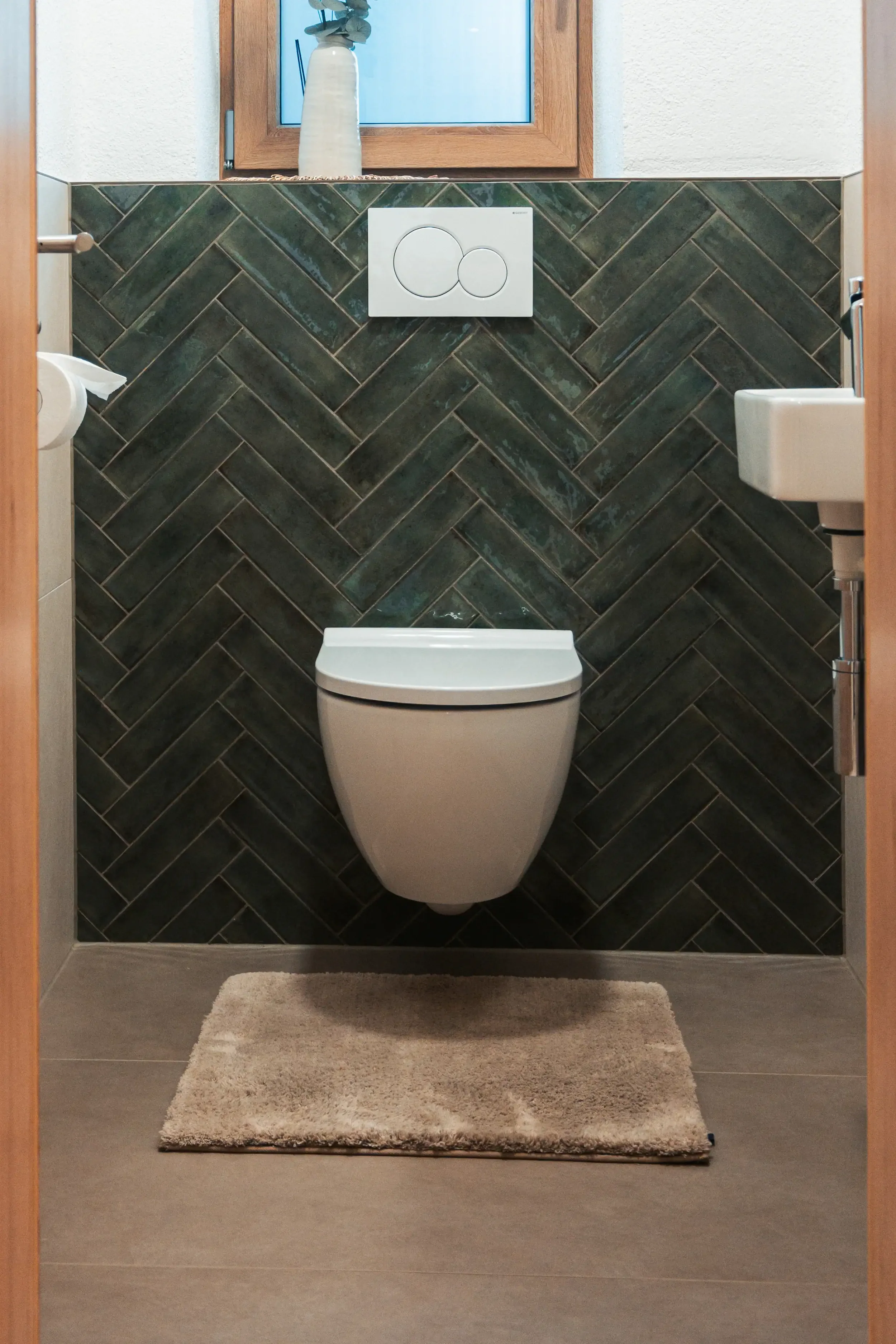 Ein Badezimmer mit einer modernen, wandhängenden Toilette, einer braunen Badematte, Fliesen im Fischgrätmuster an der Wand und einem kleinen Waschbecken. Es gibt eine Vitrine mit einer Pflanze auf der Fensterbank.