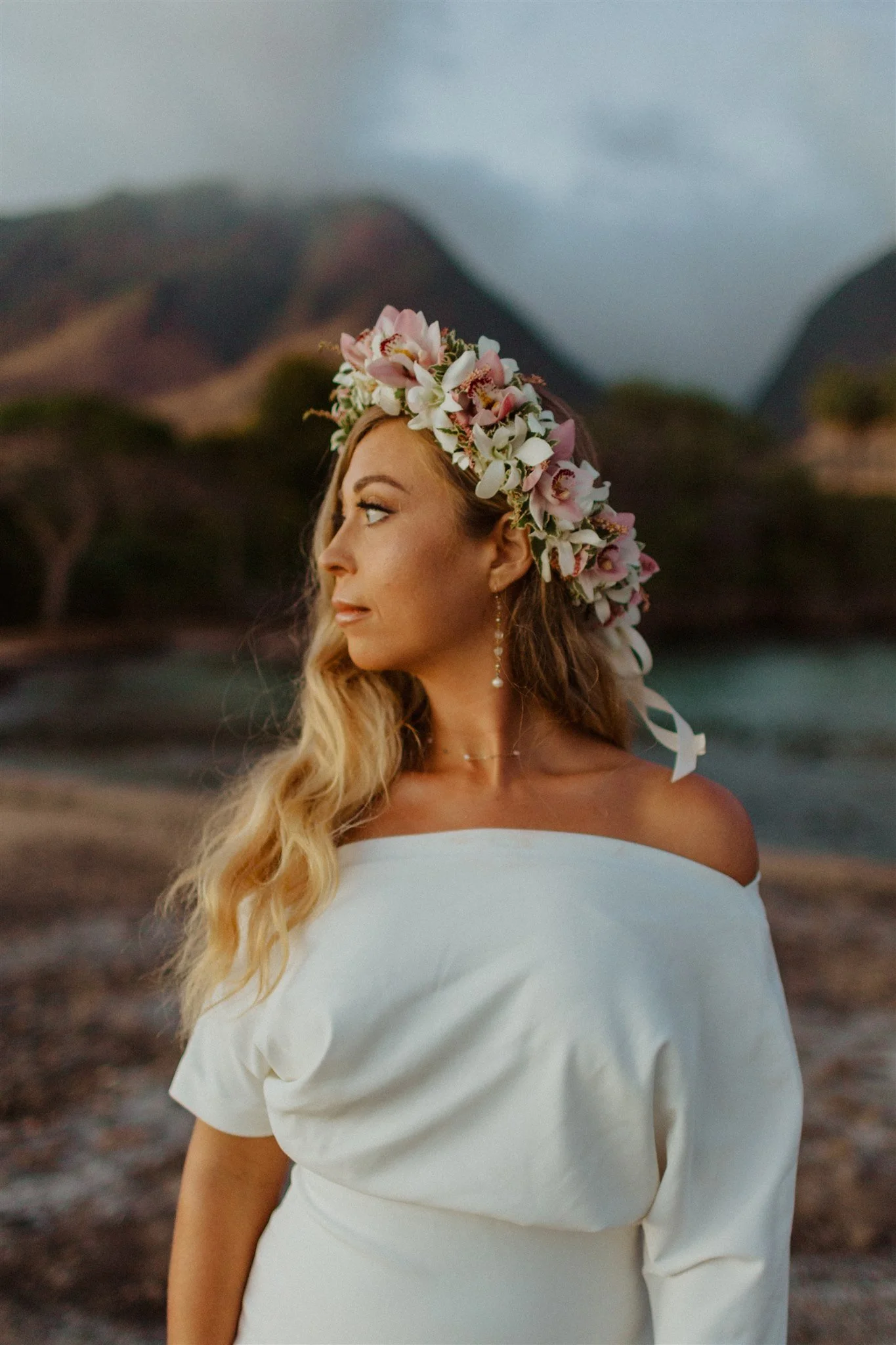 A woman with long wavy blonde hair wearing a white off-shoulder dress and a floral crown of pink and white flowers, standing outdoors near a body of water with mountains in the background.