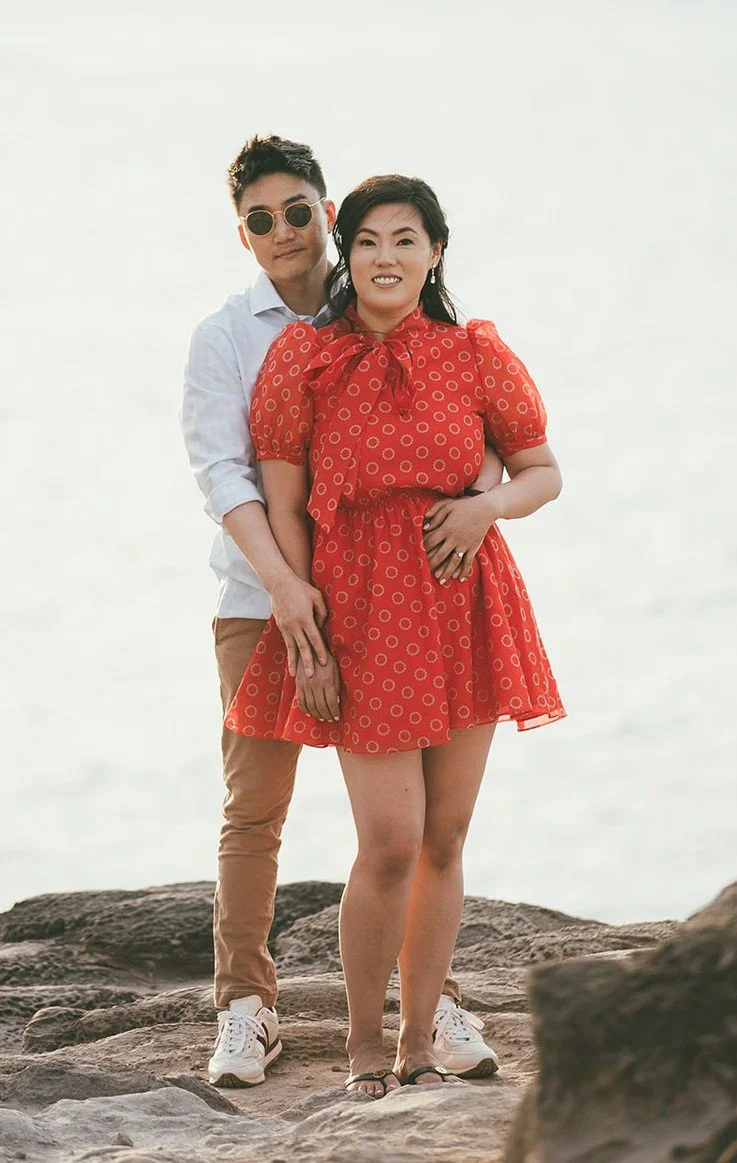A couple standing on rocks near the water, with the man behind the woman, both smiling.