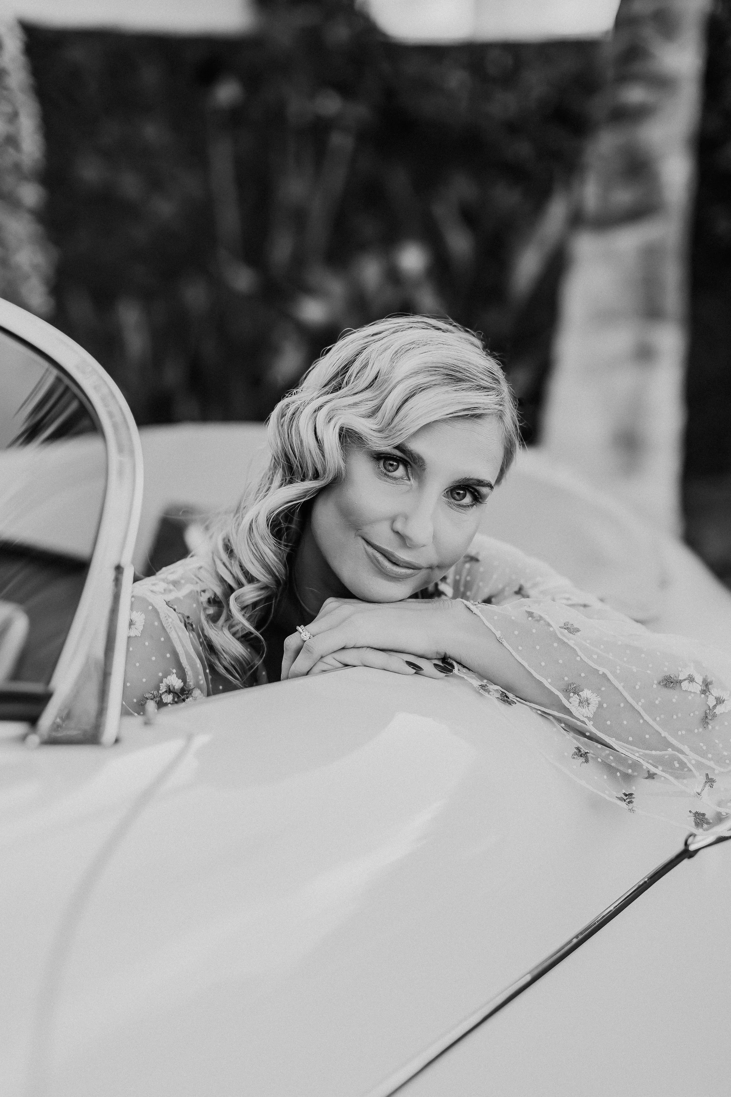 A woman with wavy blonde hair resting her chin on her hands, leaning on a vintage car hood, smiling softly at the camera.