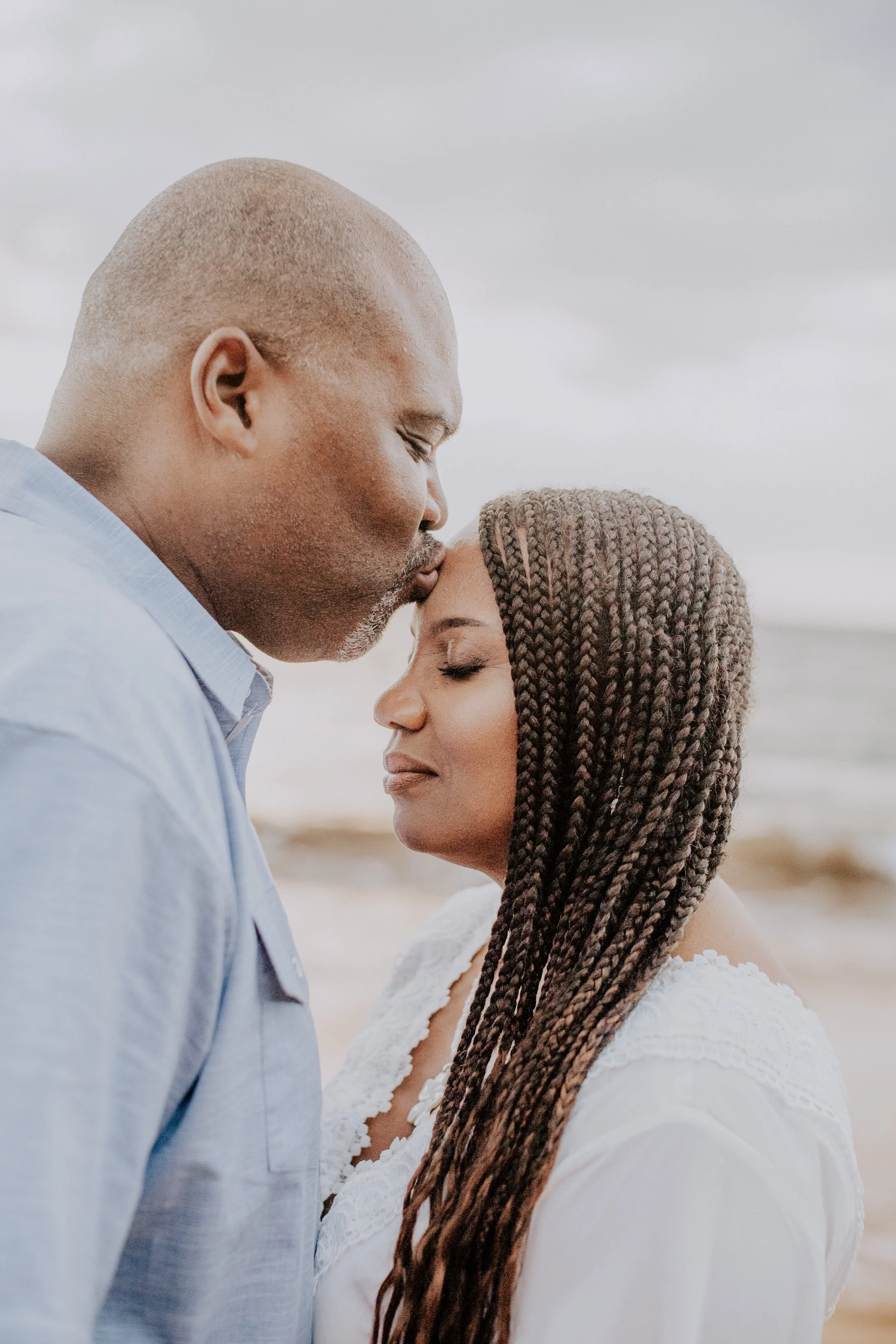 A loving couple at the beach, with the man gently kissing the woman on her forehead, both with eyes closed and peaceful expressions.