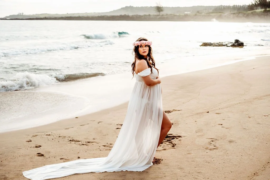 A pregnant woman standing on a sandy beach wearing a flowing white dress and a pink floral headband, with waves and a distant shoreline in the background.