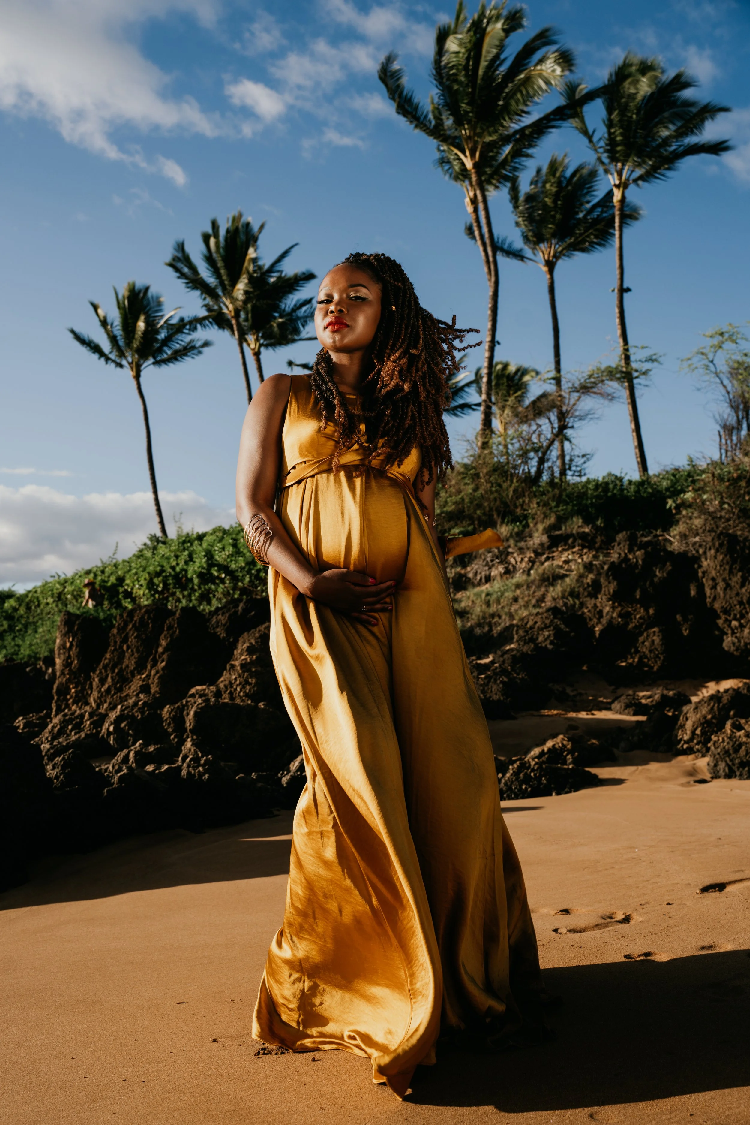 A woman in a gold dress standing on a beach with rocky terrain and tall palm trees in the background under a partly cloudy sky.