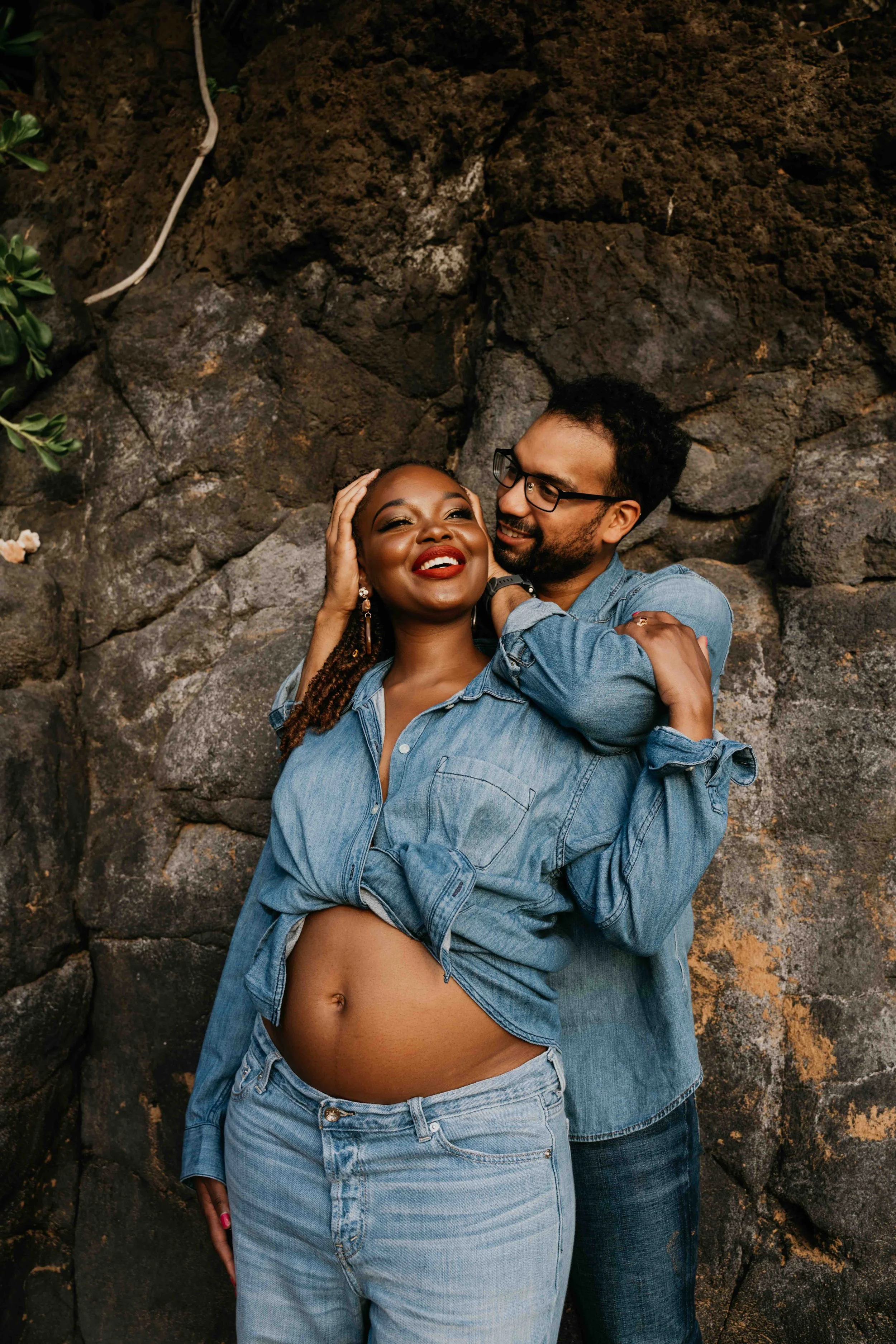 A happy couple wearing denim shirts and jeans, embracing each other against a rocky background. The woman has a big smile, visible belly button, and braided hair, while the man wears glasses and has a beard.