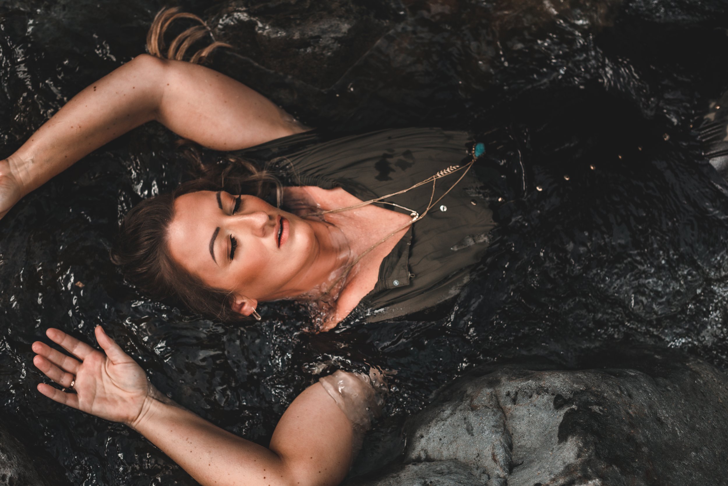 A woman with long hair floating in a dark water body, eyes closed, wearing a black top, with jewelry around her neck and ear, surrounded by rocks.