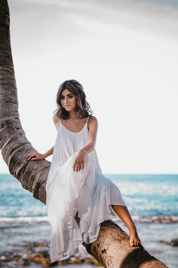 A young woman with wavy hair wearing a white flowy dress sitting on a large palm tree branch near the ocean.