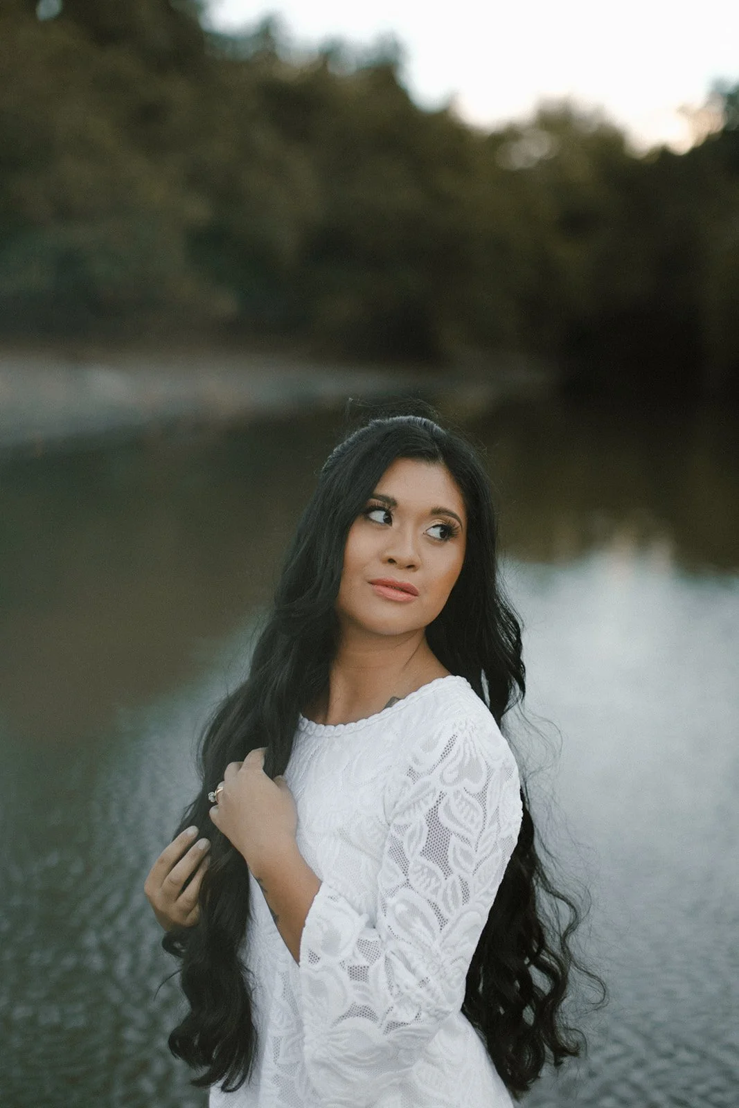 A woman with long black hair wearing a white lace dress standing near a body of water with trees in the background.