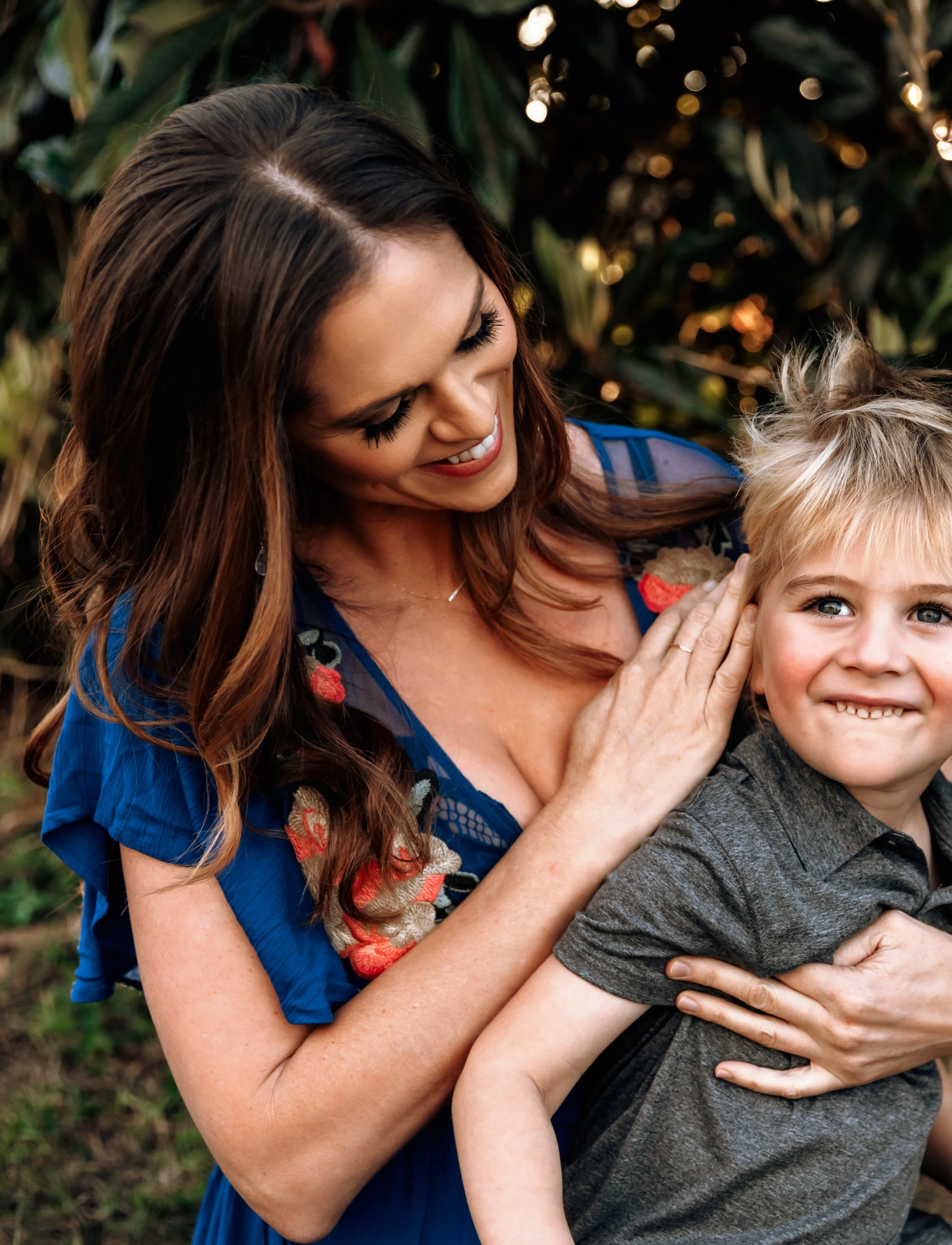 A woman with long brown hair smiling and holding a young boy with blonde hair outdoors.