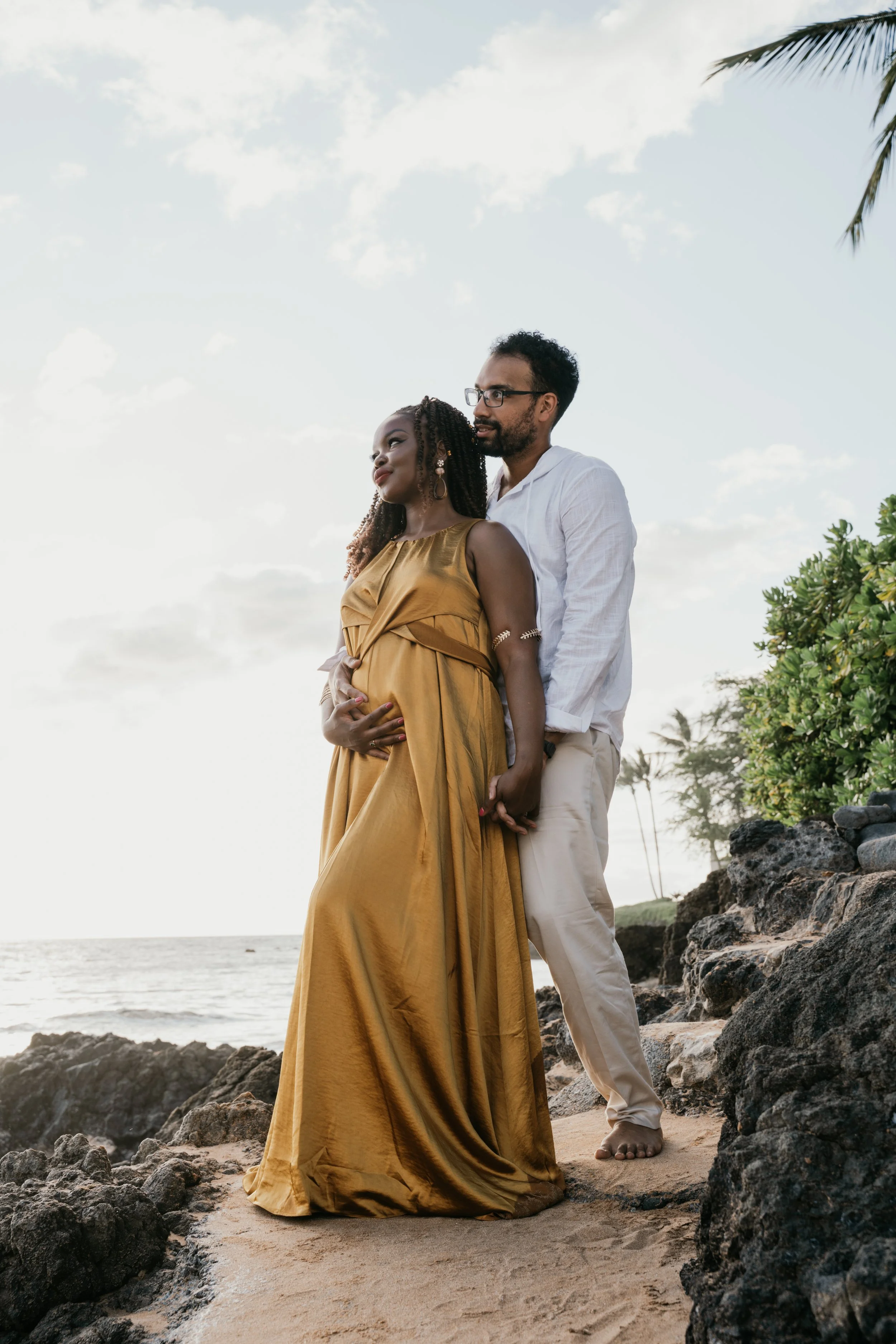 A pregnant woman in a gold dress stands on a rocky beach with her partner, who is holding her and standing behind her. The woman has braids and earrings, and they are facing a sunset or sunrise sky with the ocean in the background, surrounded by tree