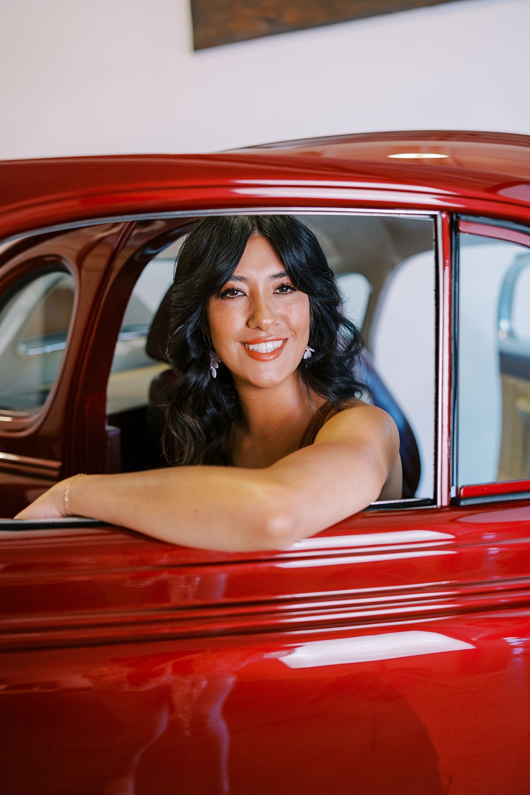 A woman with dark, wavy hair and a bright smile sitting inside a shiny red vintage car, resting her arm on the window frame.