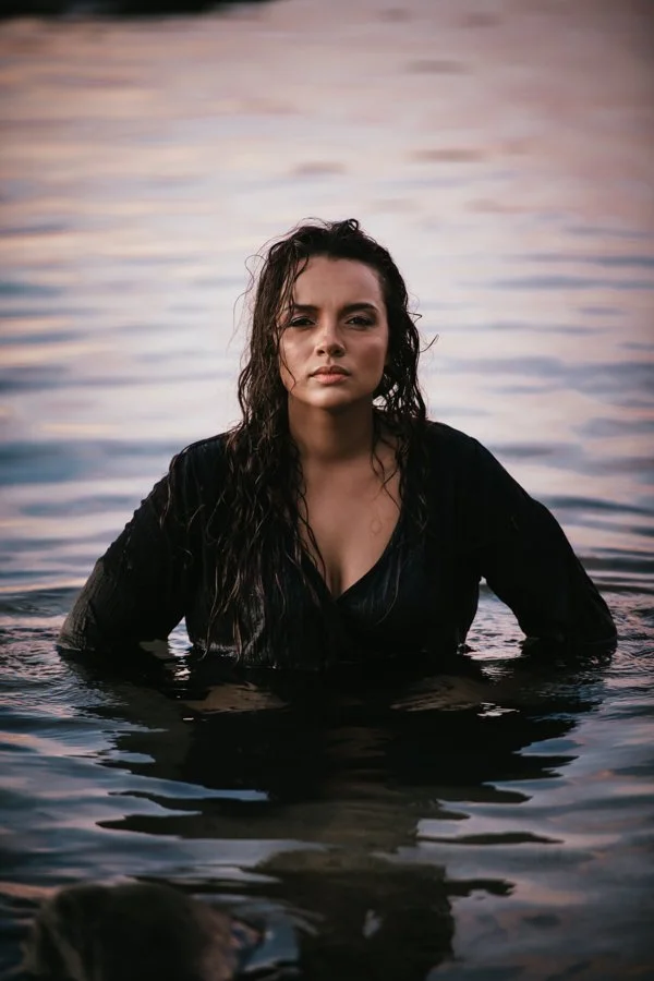 A woman with wet, wavy hair standing in a body of water, looking directly at the camera with a serious expression, during sunset or sunrise.