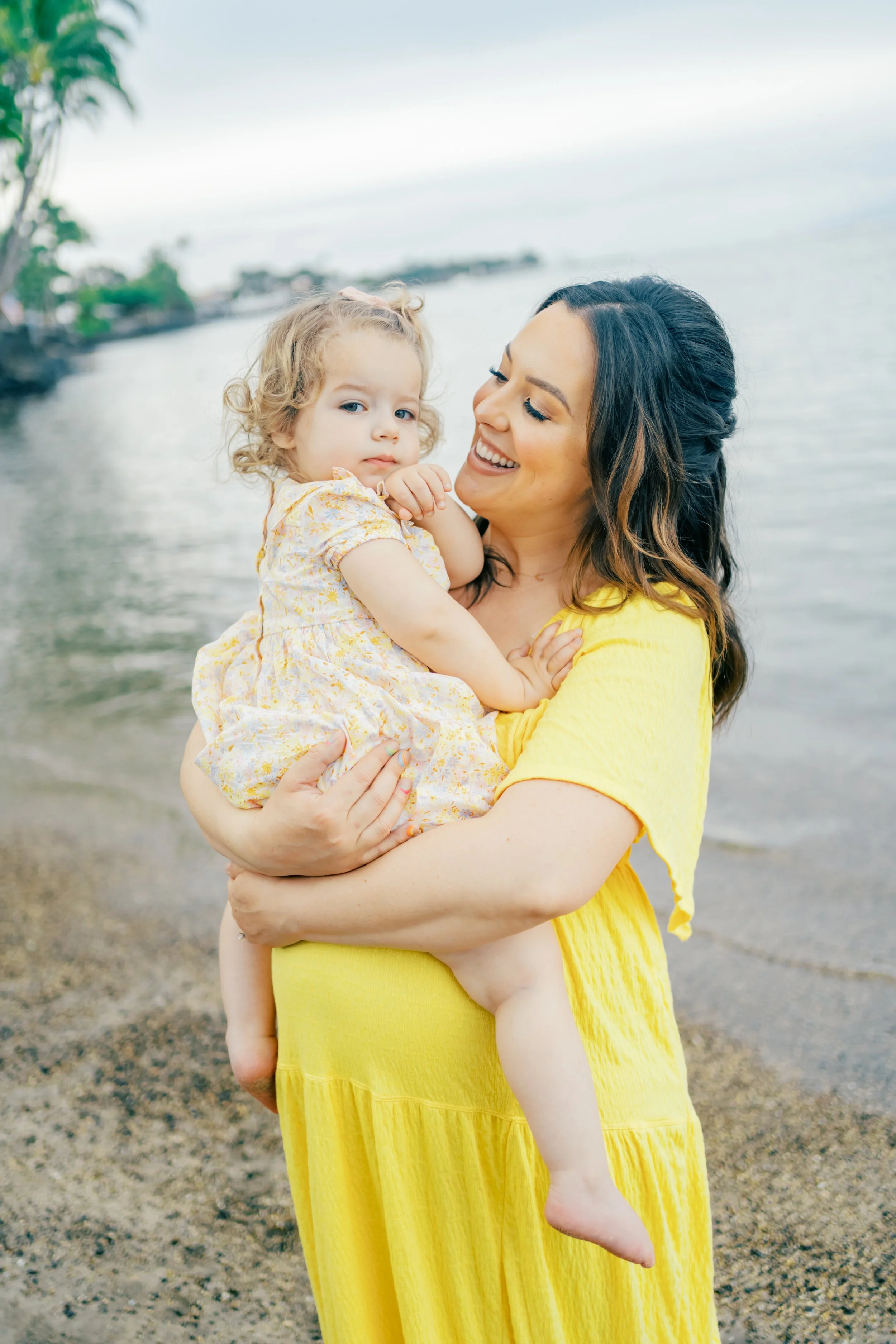 A woman holding a young girl by the water at a beach, smiling at the girl who is looking to the camera.