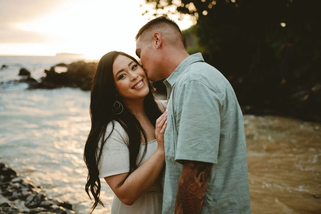 A couple at the beach, the woman smiling at the camera as the man kisses her cheek, sunset in the background.
