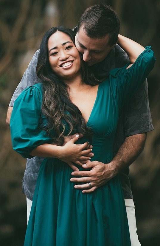 A smiling woman in a teal dress being embraced from behind by a man, both appearing happy and close together outdoors.