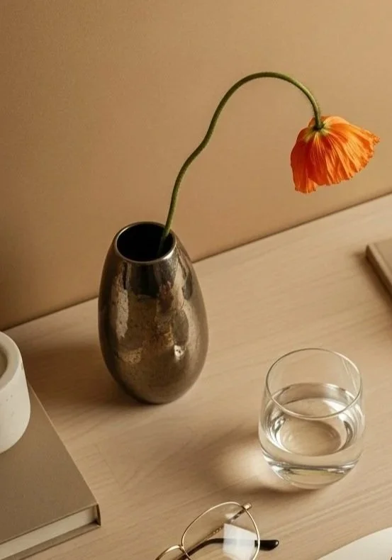 A single orange flower in a metallic vase, with a glass of water and a pair of glasses on a wooden table.