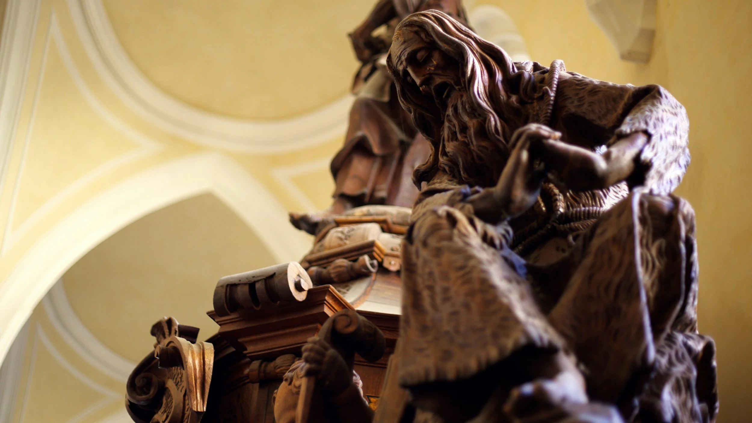 Close-up of a wooden statue of a seated man with a beard, holding an open book, against a blurred background of yellow walls and architectural details.