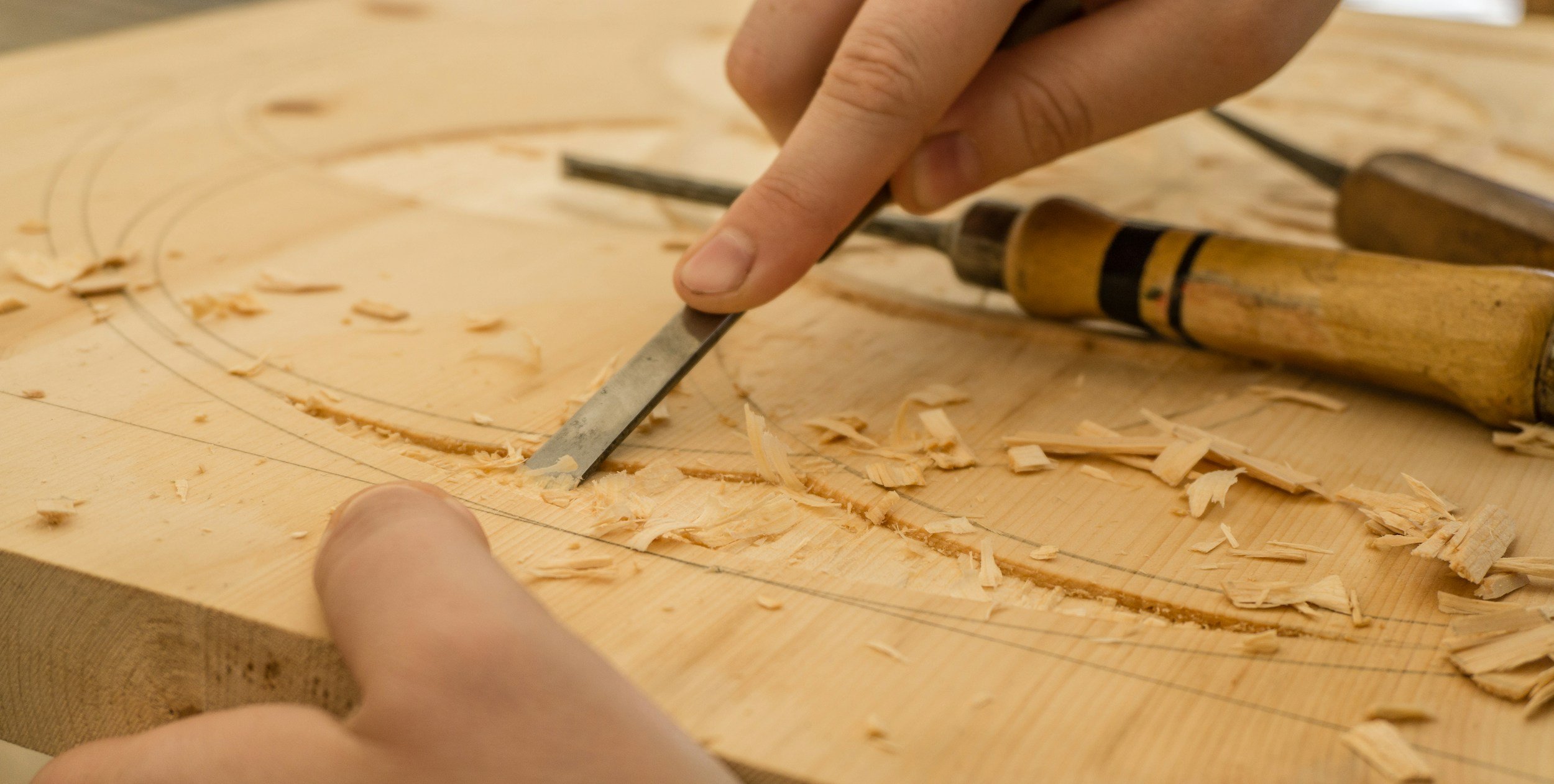 A person is carving wood with a chisel and mallet on a wooden workbench. Wood shavings are scattered around.
