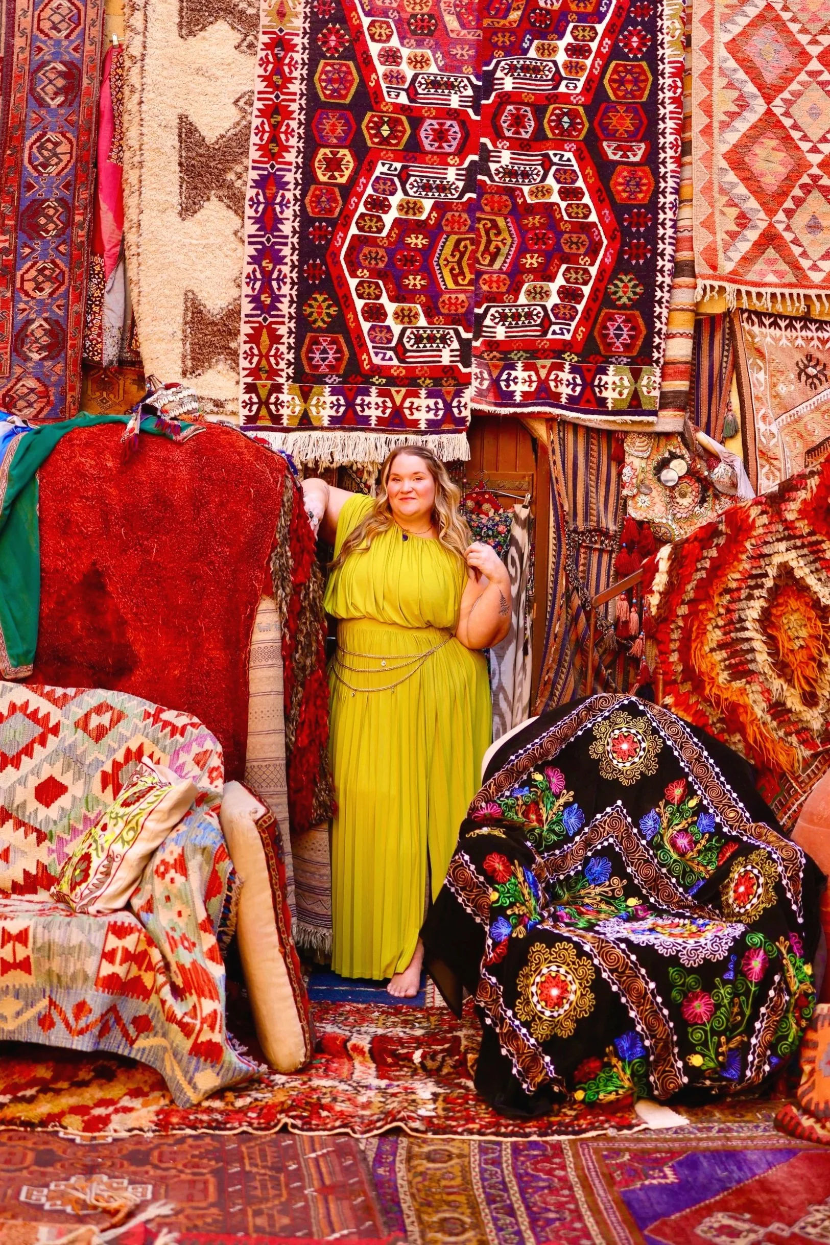 A woman in a bright yellow-green dress standing amidst a display of colorful, patterned rugs and textiles hanging on the wall and draped over furniture in a rug shop.