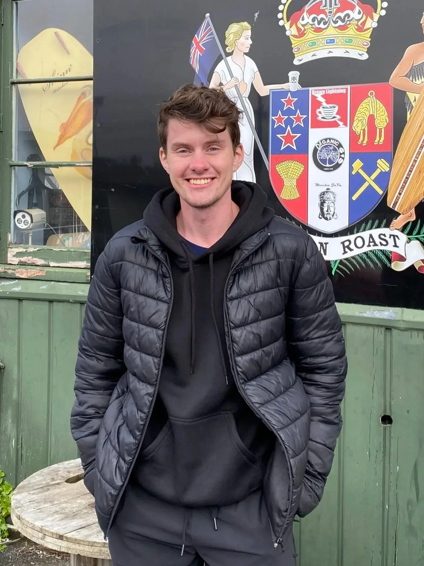 A young man with short brown hair wearing a black puffer jacket and black hoodie smiling and standing outdoors in front of a colorful shield with various symbols, a woman holding an Australian flag, and a crown, with a green wooden wall behind him.