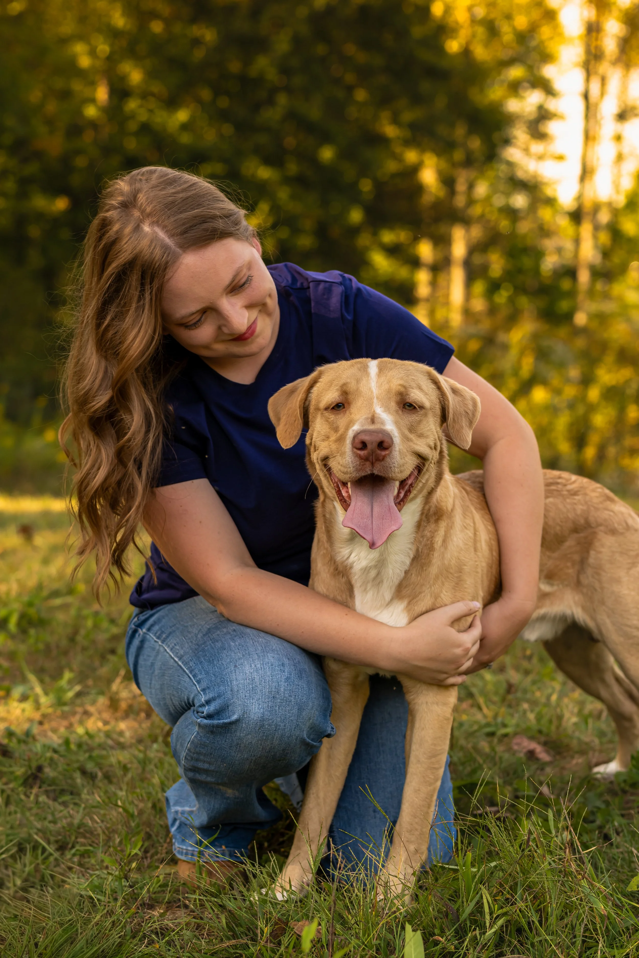 Woman in blue shirt kneeling outdoors with a happy, tan dog in a grassy area with trees and sunset background