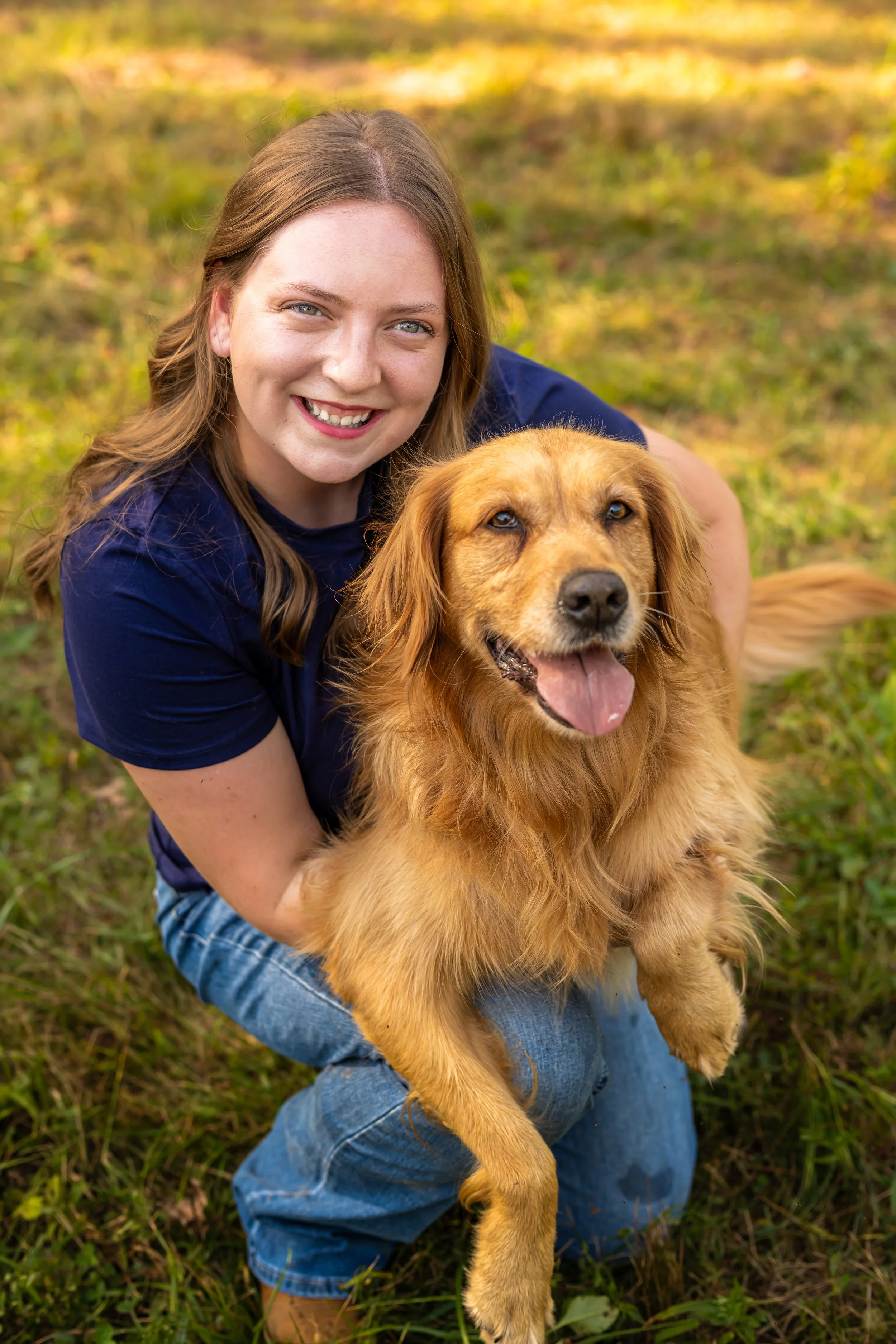 A young woman with long brown hair, wearing a navy blue T-shirt, holding a golden retriever dog outdoors on grass in a park, both smiling.