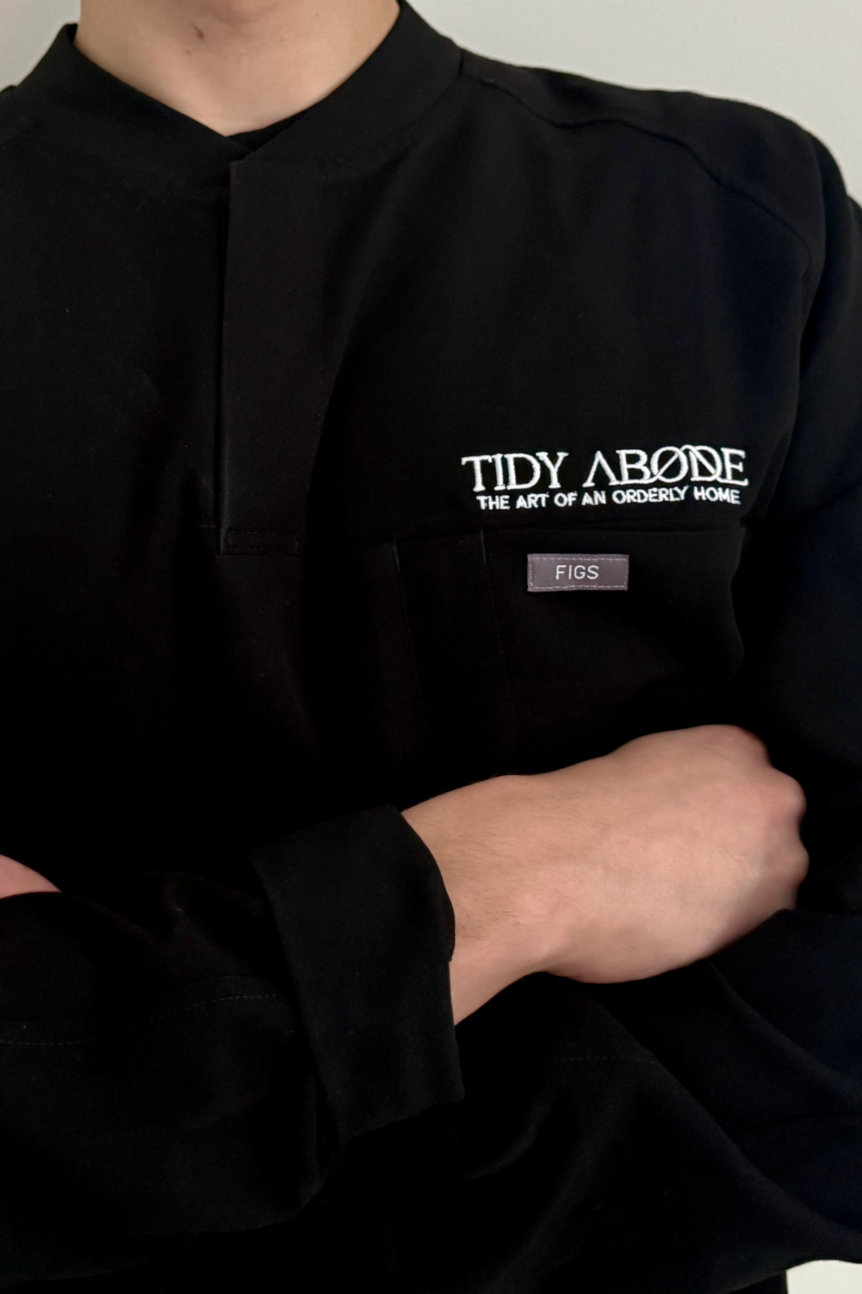 Close-up of a person wearing a black work jacket with the text 'TIDY ABODE THE ART OF AN ORDERLY HOME' embroidered on the chest and a small label reading 'FIGS' on the pocket.