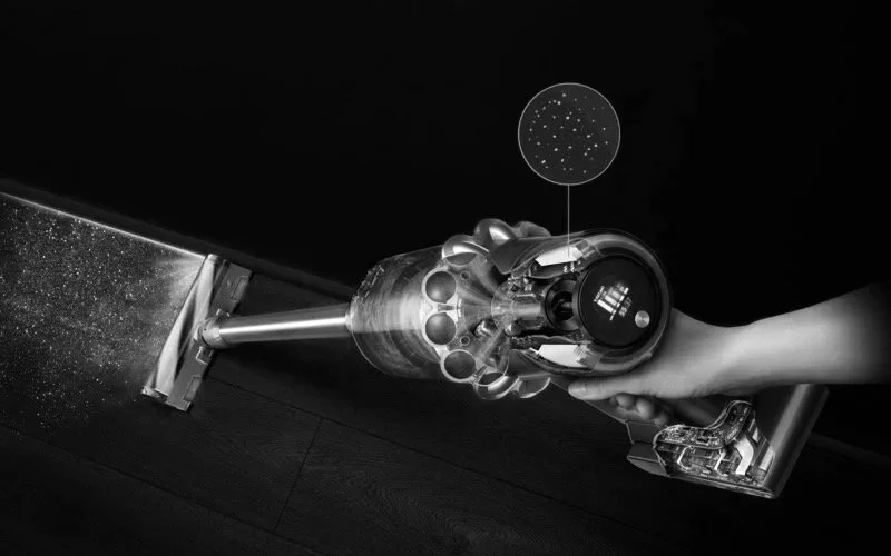 A person holding a cordless vacuum cleaner with a transparent dust canister, cleaning a dark hardwood floor, with a digital star map overlay in the background.