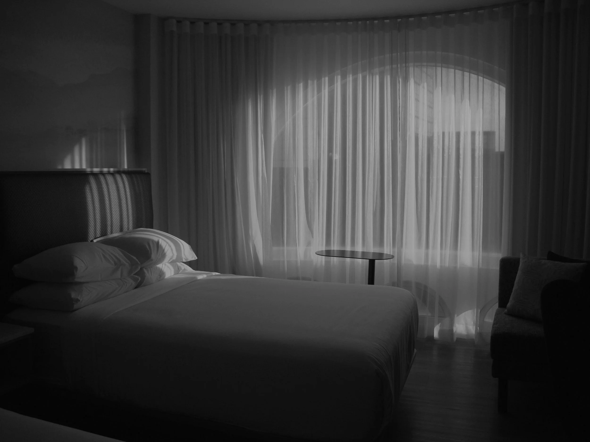 A black and white photograph of a hotel room featuring a made bed with pillows, a bedside table, a small round table near a window with curtains, and an armchair with a cushion, with natural light filtering through the window.