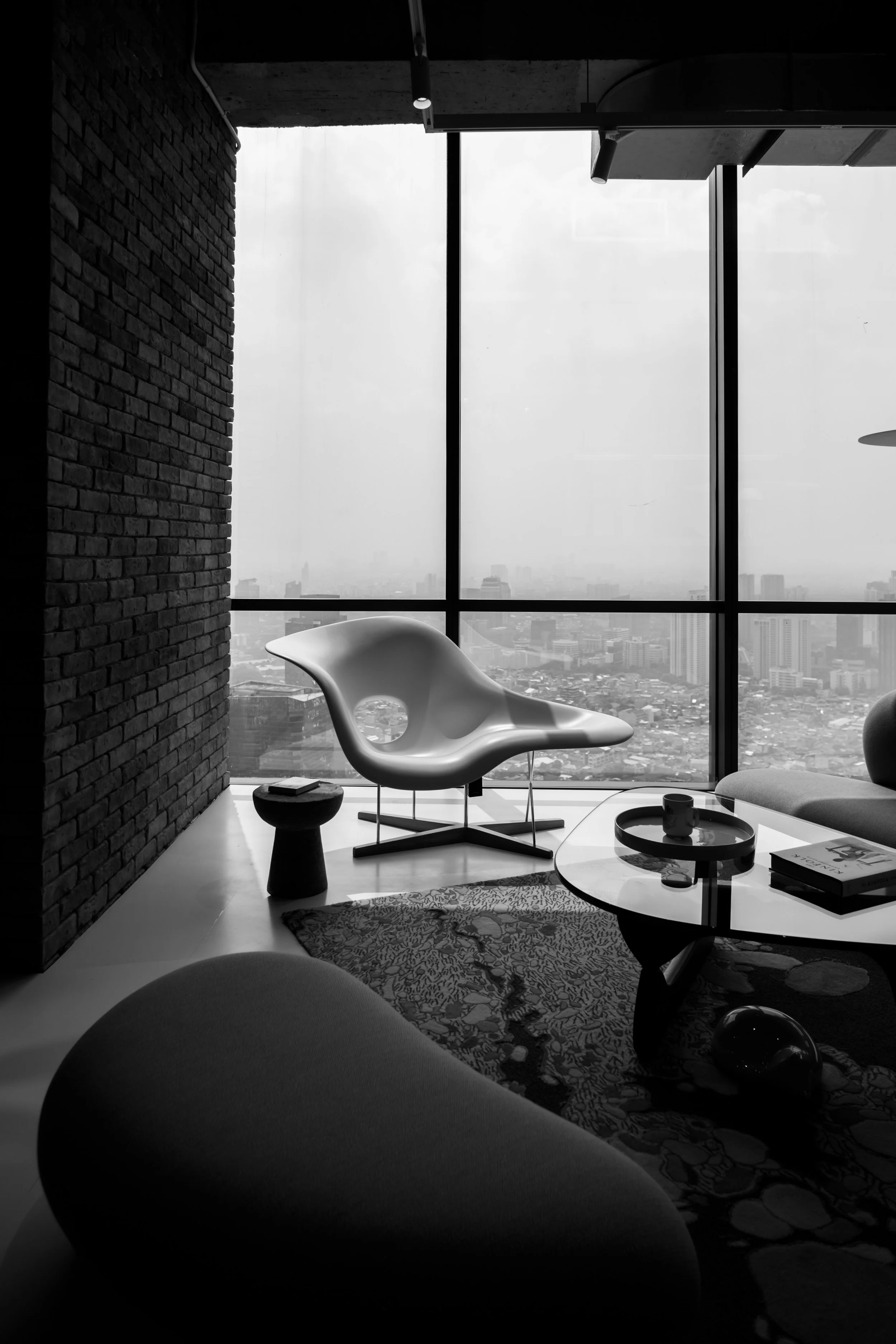 Modern living room with a large floor-to-ceiling window overlooking a cityscape, featuring a white lounge chair, a small side table, a round coffee table with books and a mug, and a patterned rug.