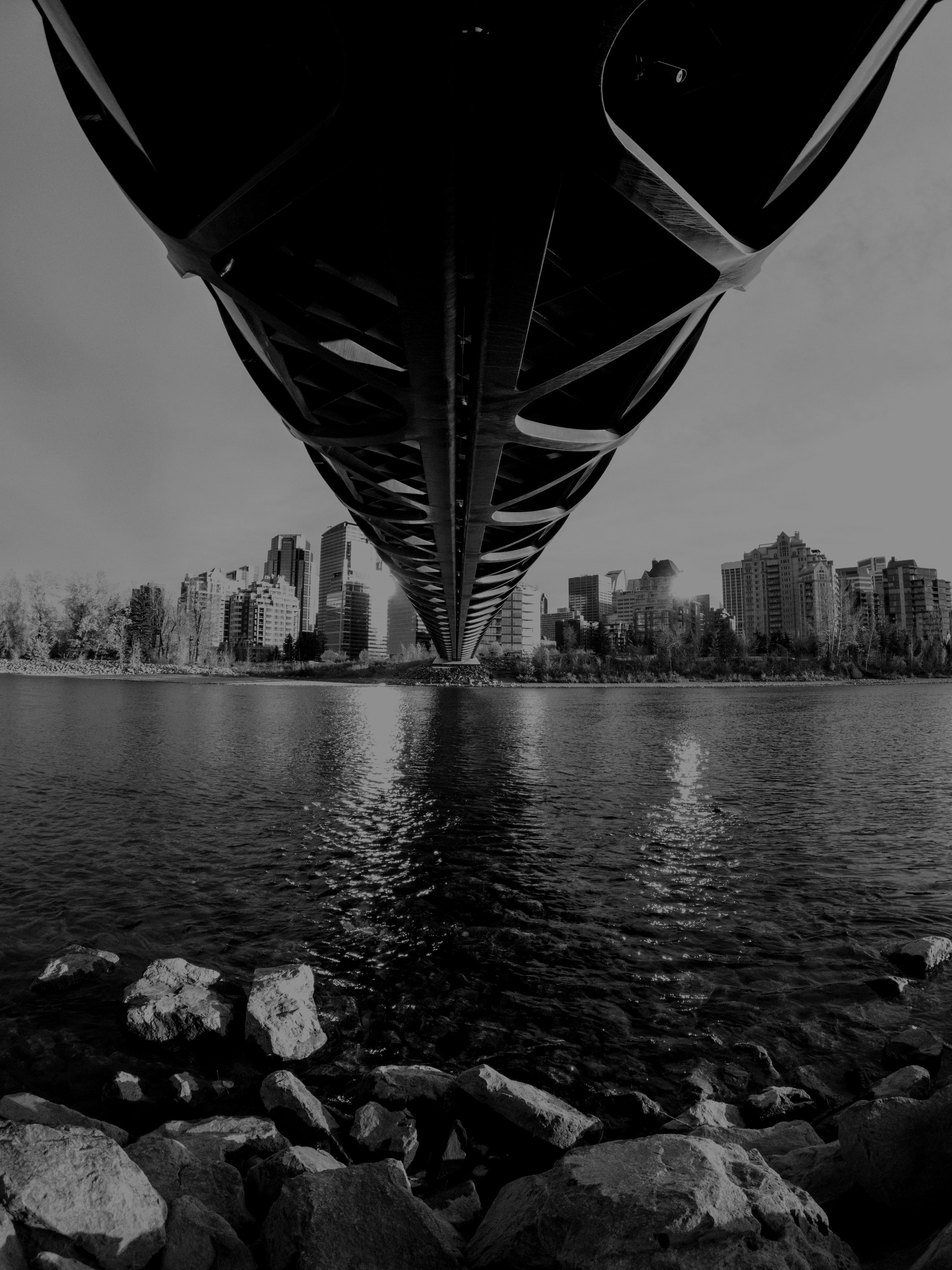 Low-angle view of a bridge over a body of water with a city skyline in the background, in black and white.