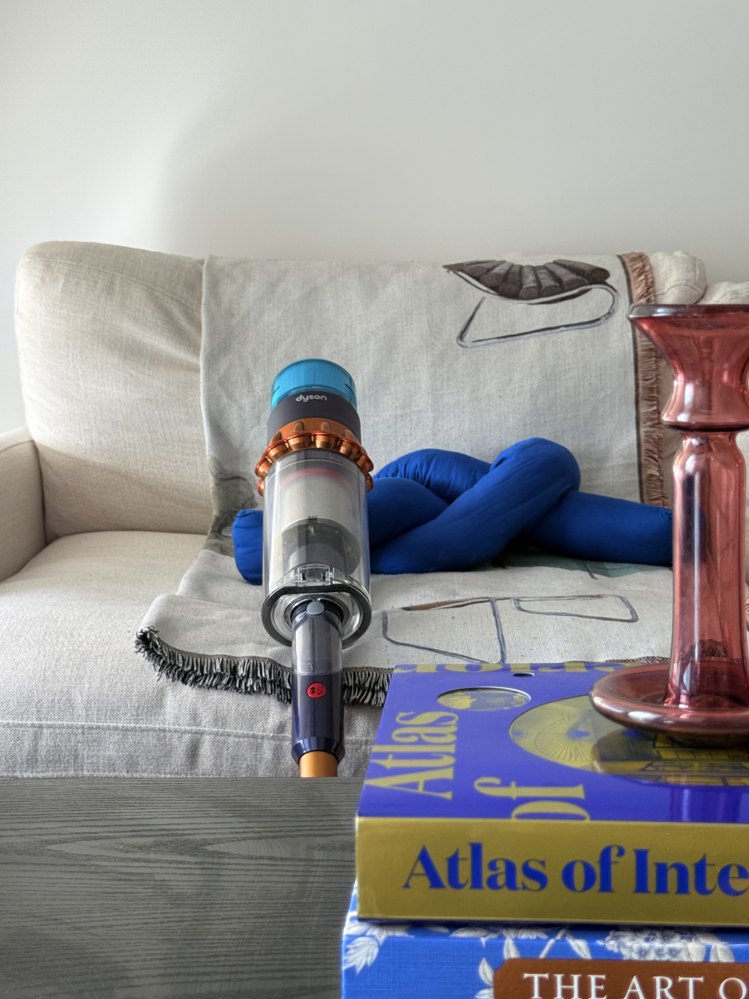 A living room with a beige couch, a gray blanket featuring a snake and other patterns, a blue plush toy, a purple Dyson vacuum cleaner on the couch, and a table with a blue 'Atlas of Interior' book and a red glass vase.