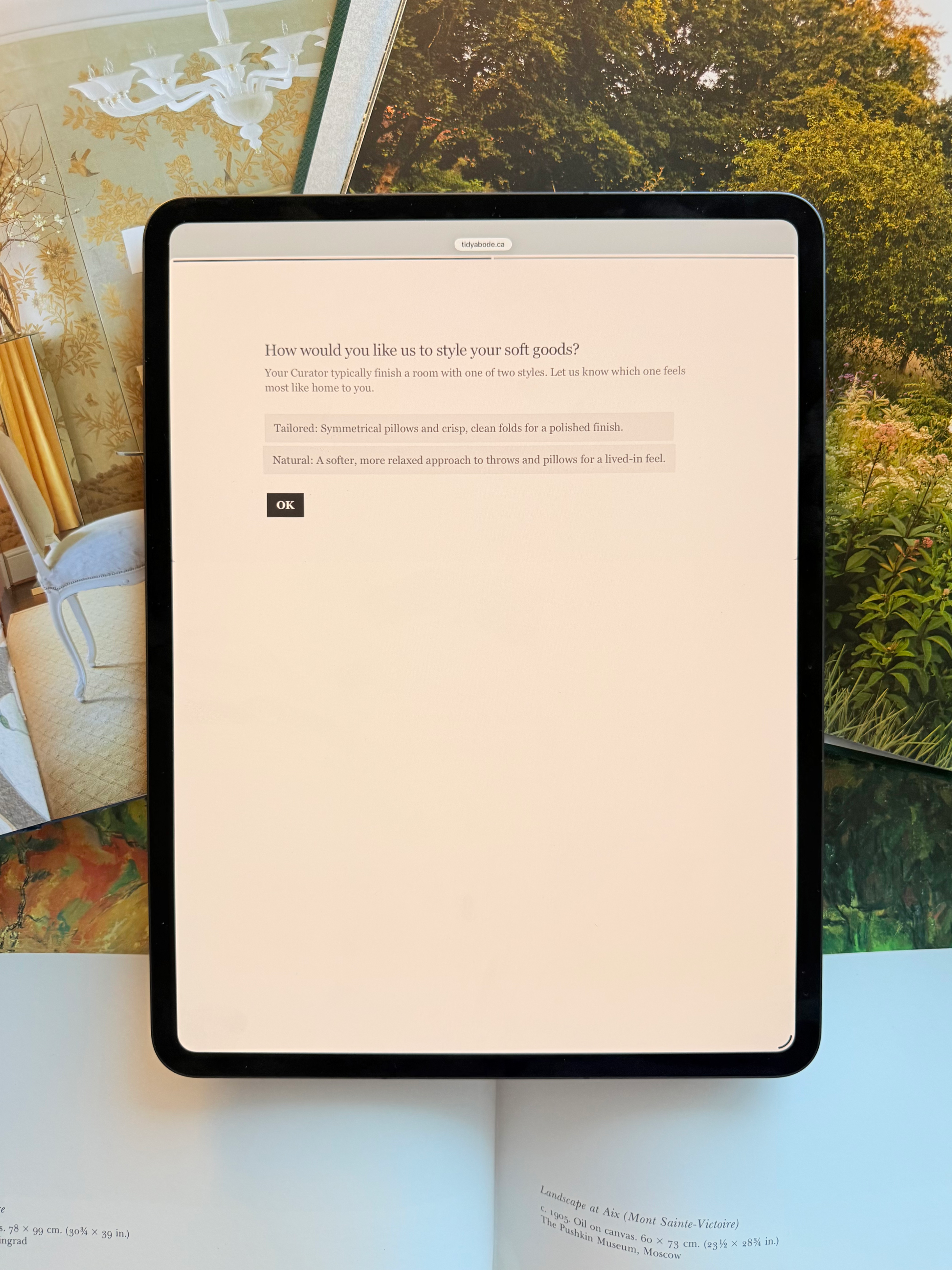 Tablet displaying a webpage about styling soft goods, with a background of outdoor trees and various open books or magazines with decorative images and text.
