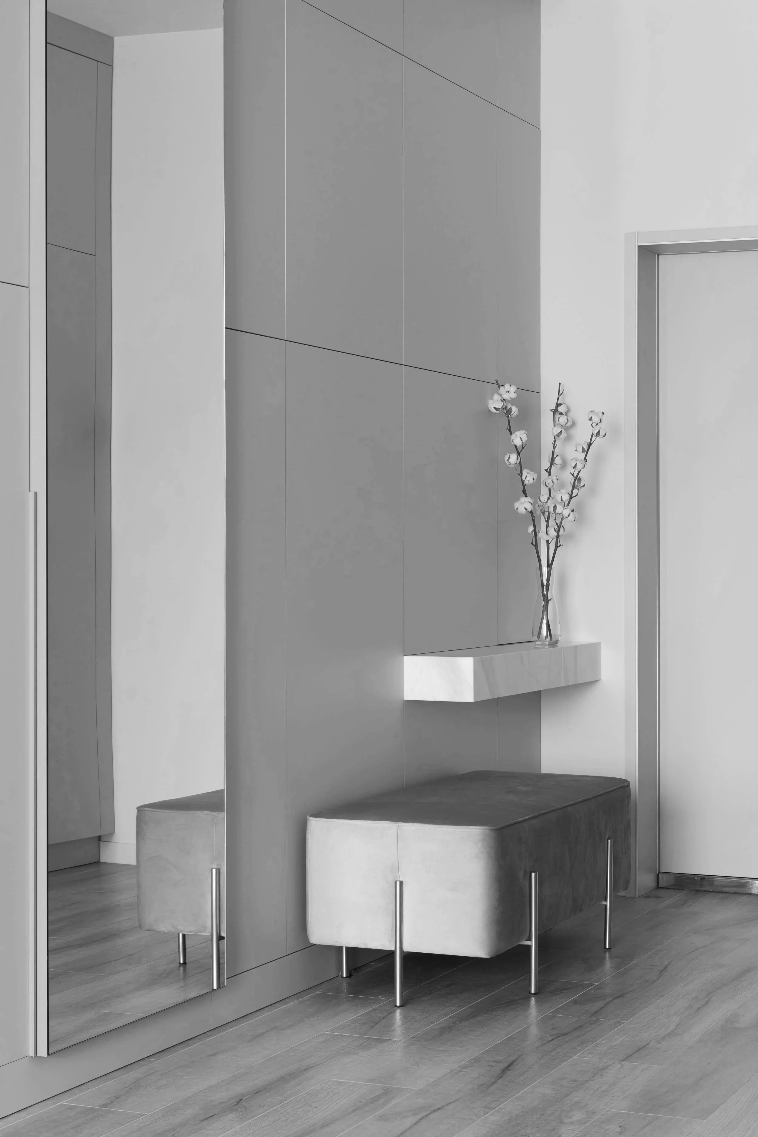 A black and white interior scene featuring a wall with mounted panels, a marble shelf with a glass vase containing cotton branches, a cushioned bench with metal legs, and a floor with wooden planks.