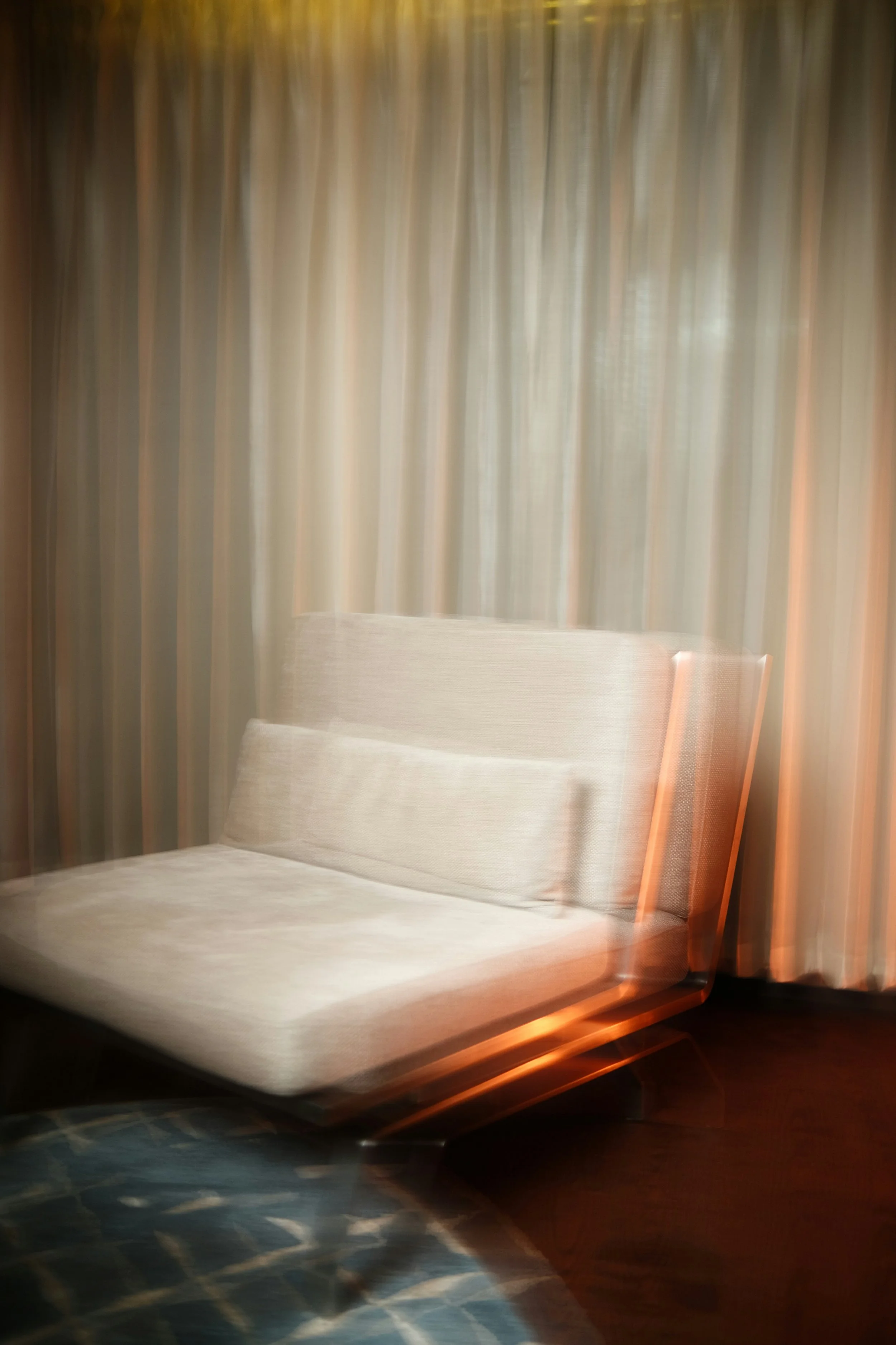 A blurred image of a beige armchair in front of beige curtains, with a dark wooden floor and a partially visible patterned fabric in the foreground.