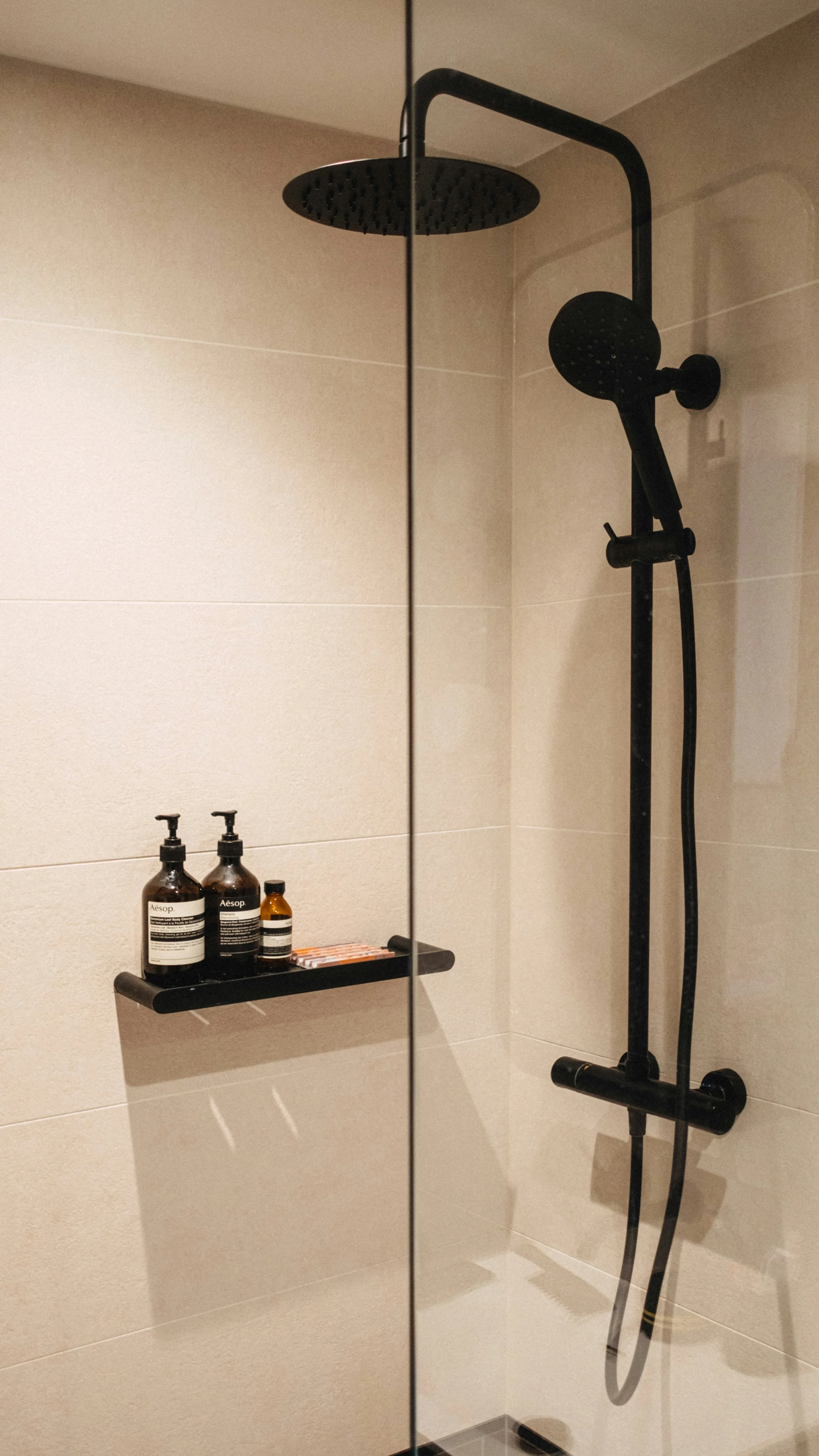 A black showerhead and handheld shower attached to a wall with beige tiles, a mirror reflecting part of the shower, and a black shelf holding bottles of soap and toiletries.