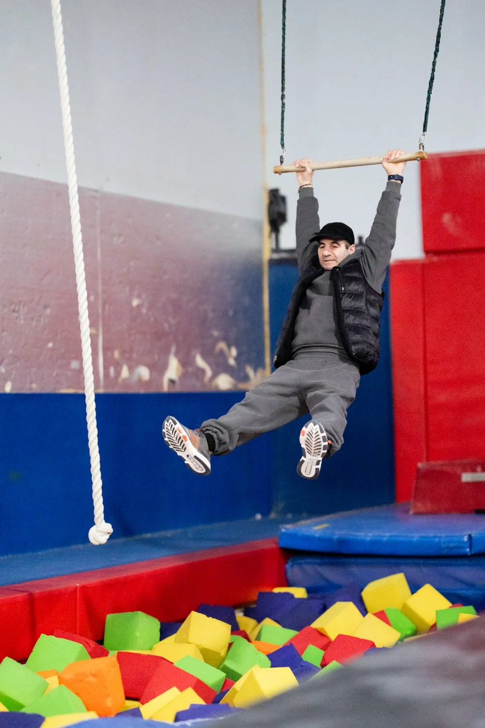 A man in gray pants, a dark vest, gray sweater, and black cap is swinging on a trapeze above a colorful foam pit at an indoor trampoline park.