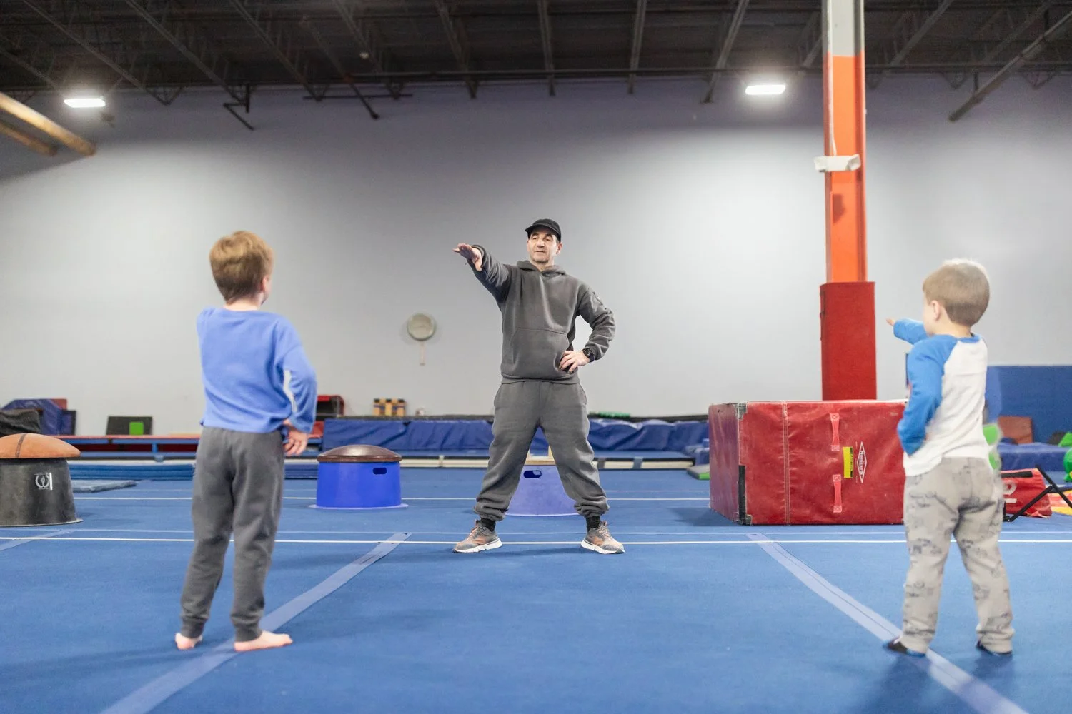 An adult male instructor demonstrating a physical exercise or stretch to two young boys in a gymnasium. The boys are standing on a blue gym floor, facing the instructor, who is gesturing with his arm. The gym contains various equipment, such as padde