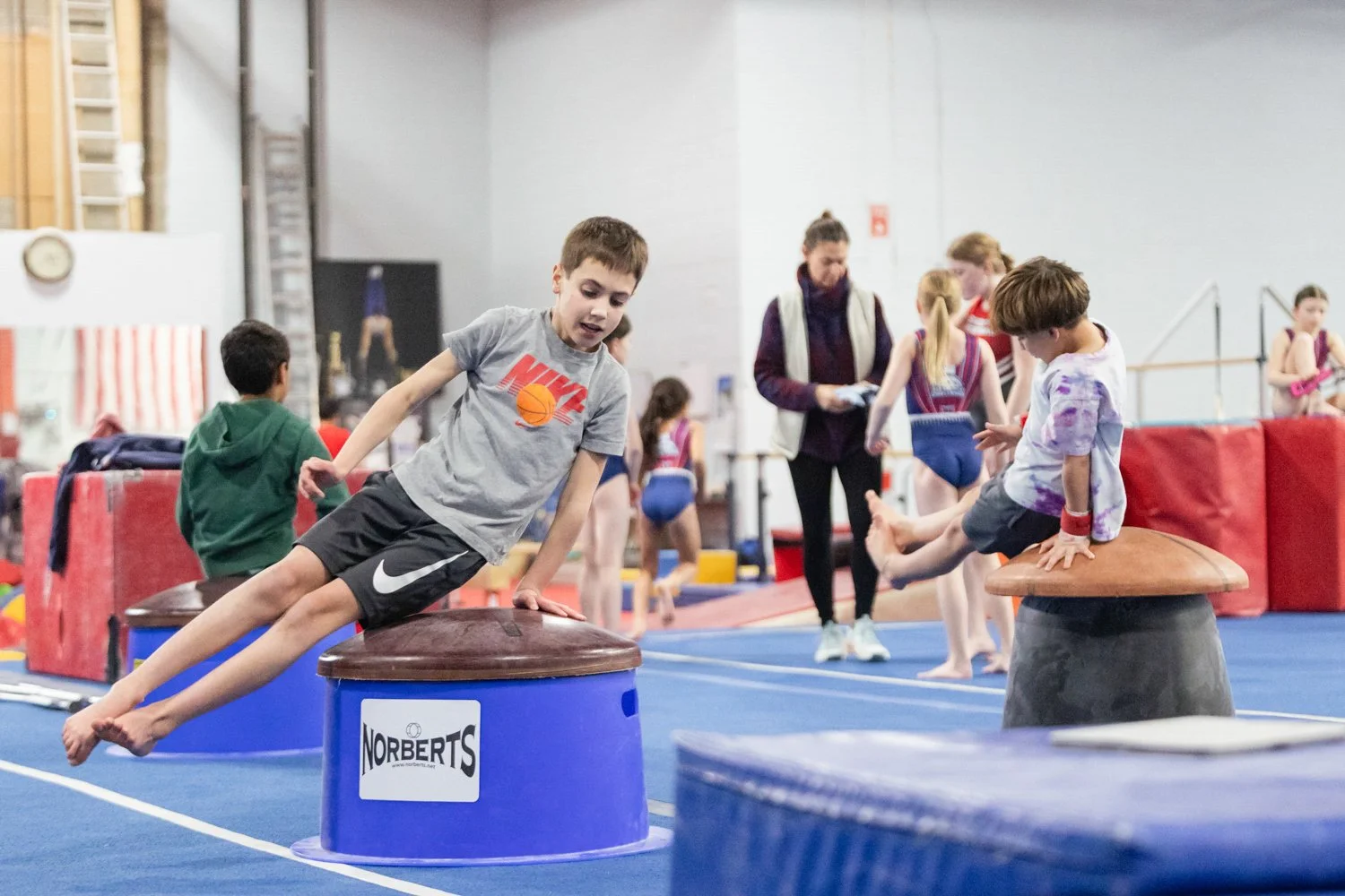 Kids participating in gymnastics activities in a gymnasium, with some kids jumping off equipment and others waiting or observing, overseen by an instructor.
