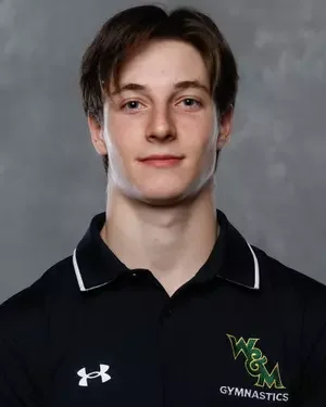 Young man with brown hair wearing a black polo shirt with a gymnastic team logo and Under Armour logo, standing against a gray background.