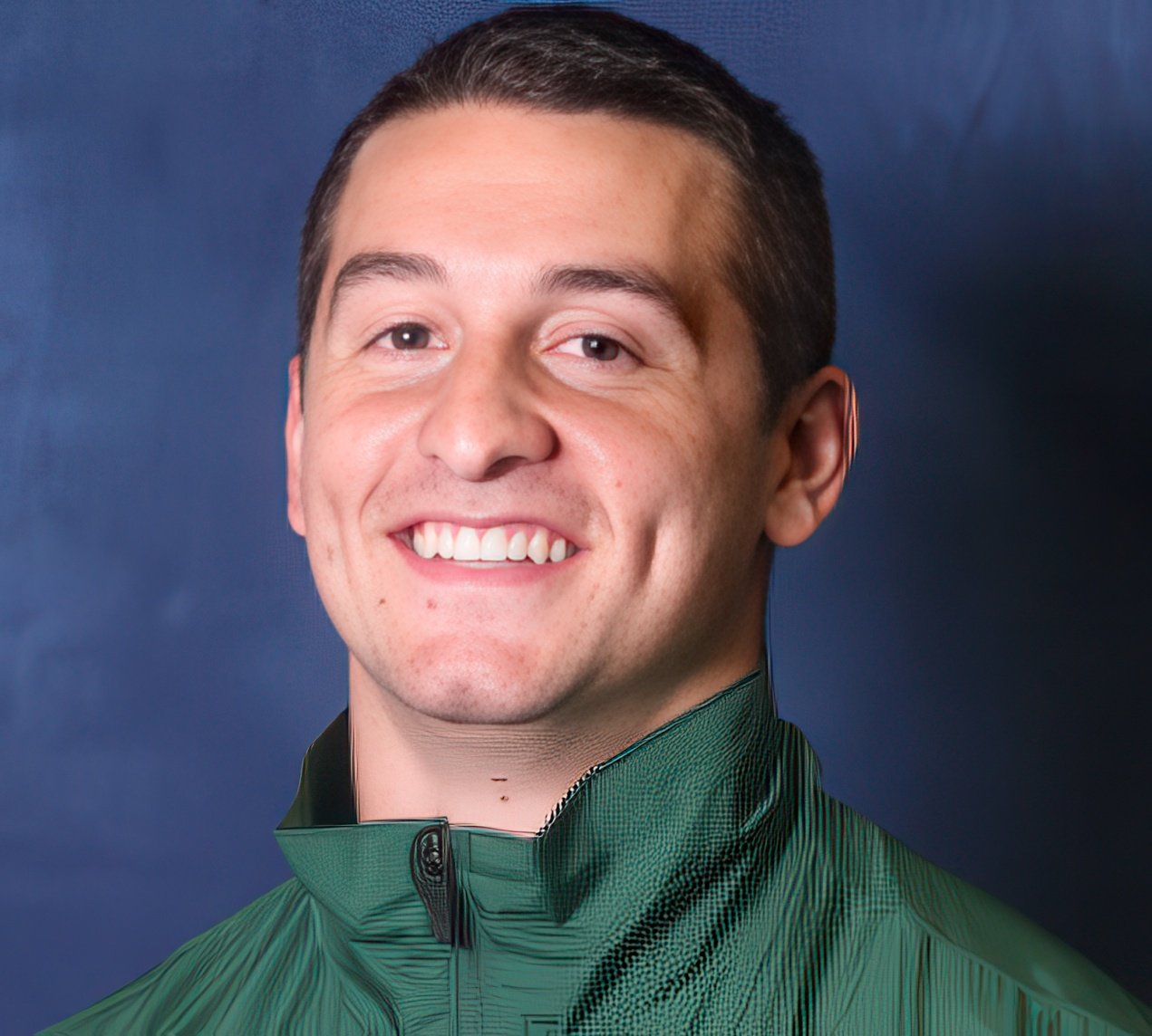 Portrait of a smiling young man with short dark hair wearing a green jacket against a blue background.