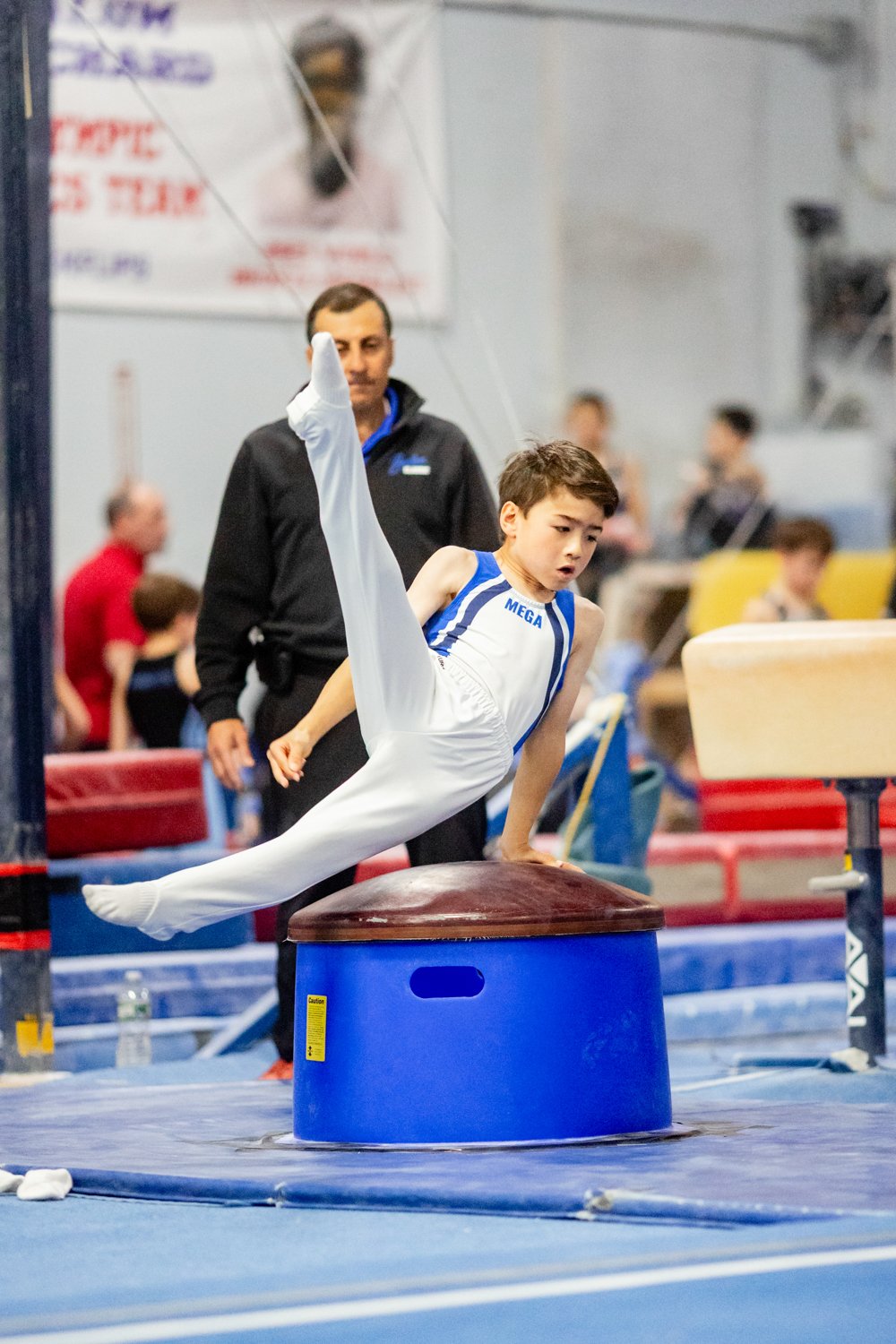 A young boy in a white and blue gymnastic uniform performing a vault on a large round mat in a gymnastics training facility, with a coach or judge observing in the background.
