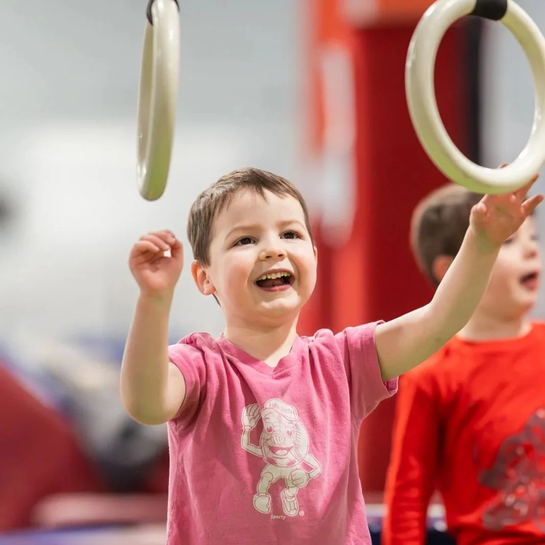 A young boy in a pink shirt smiling and reaching up towards gymnastic rings.